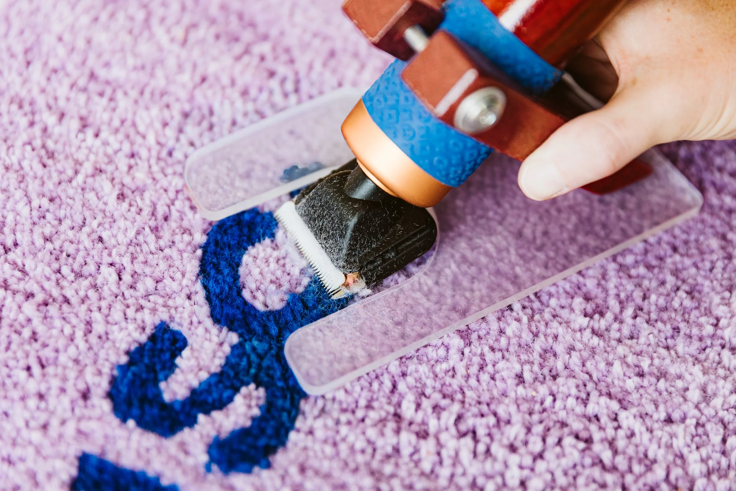 Nahaufnahme einer Hand, die ein Rasiermesser auf einem pinken Teppich hält, neben einem Schneidebrett mit blauer, schwarzer, roter und kupferfarbener Dekoration.