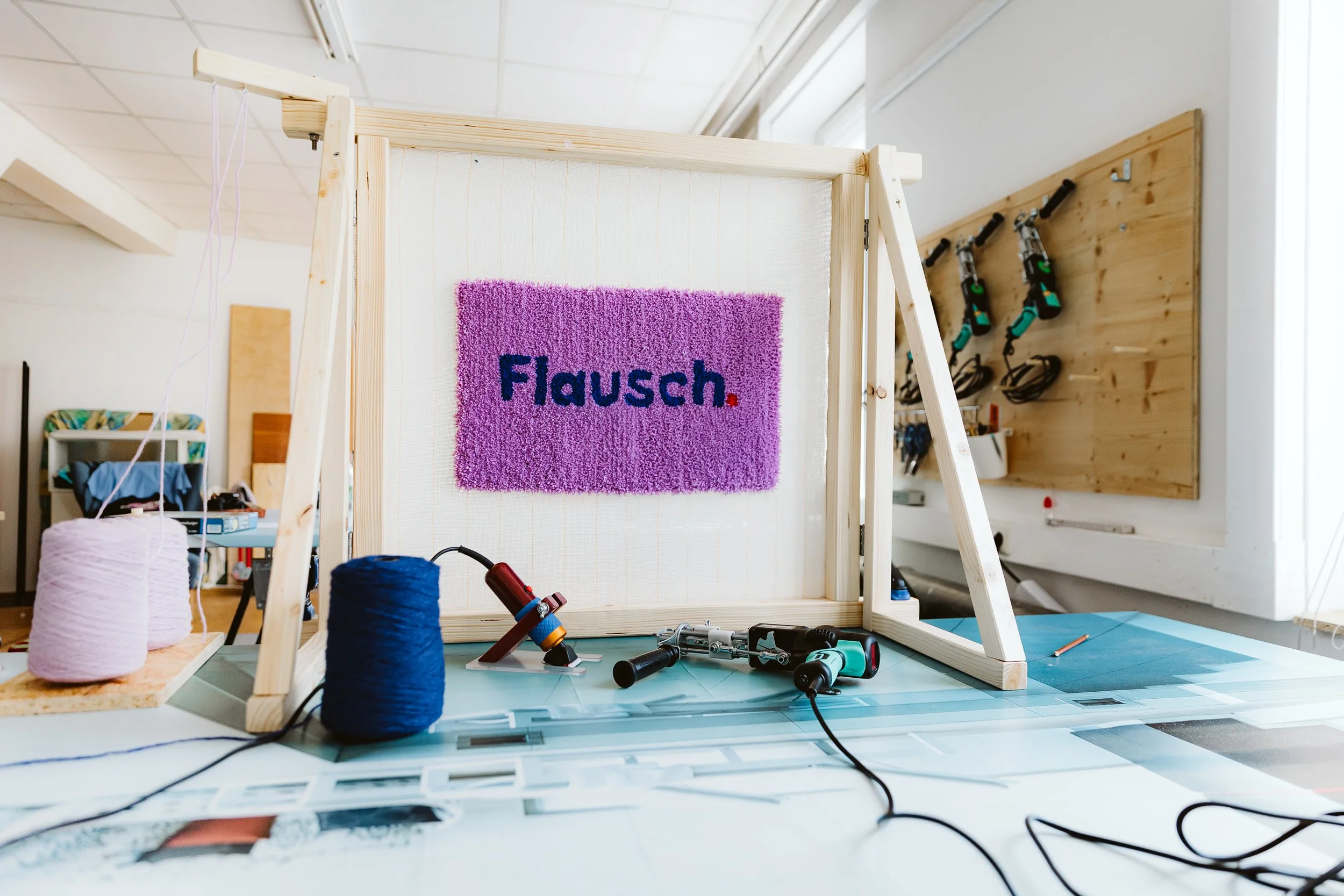 Blick auf eine Werkbank mit Nähzubehör, darunter Knäuel von Faden und Werkzeug, im Hintergrund ein Holzständer mit einem Farbmatten-Teppich, der auf einen Schriftzug mit der deutschen Bezeichnung 'Flausch' zeigt.