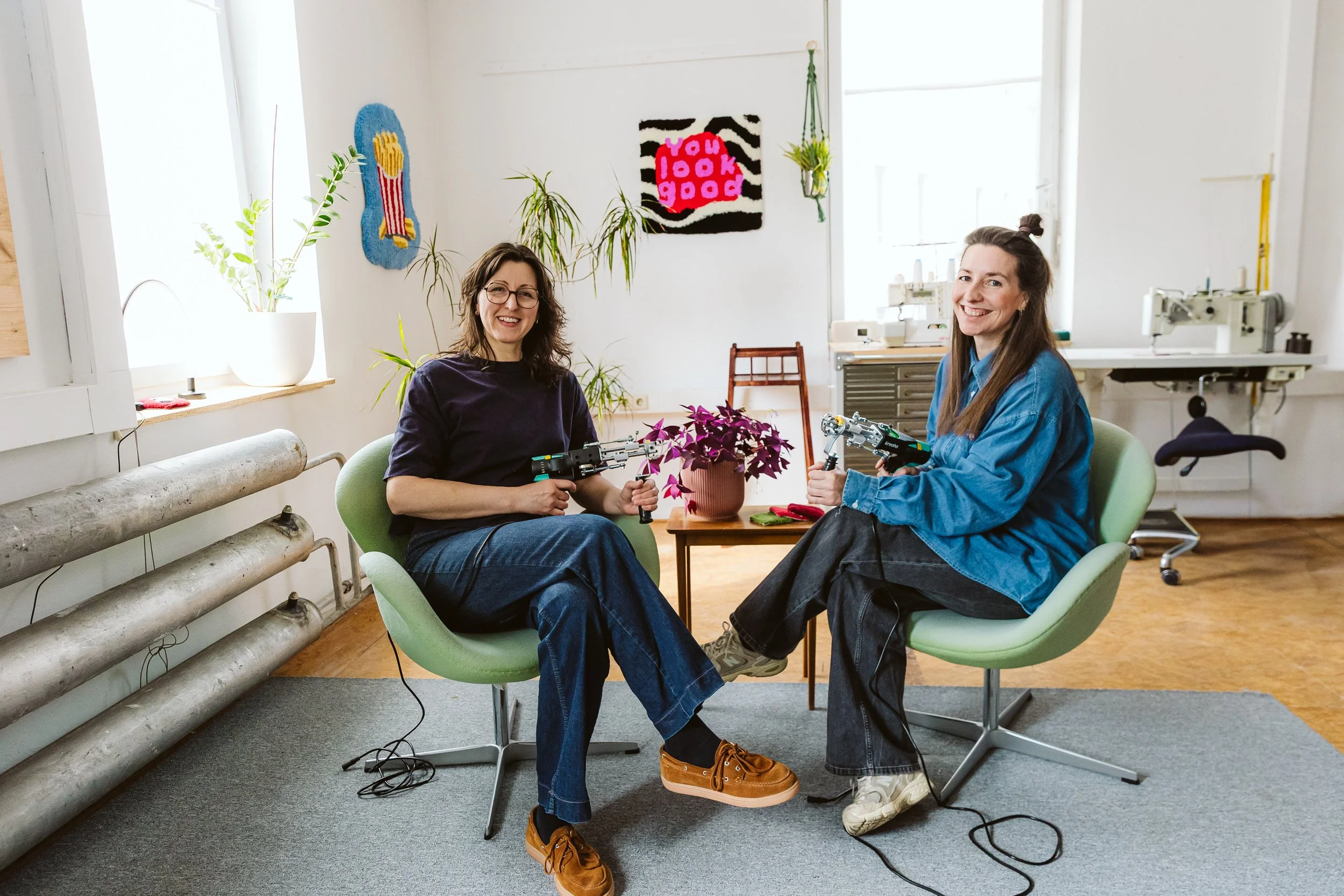 Zwei Frauen sitzen in einem Raum mit weißen Wänden und großen Fenstern, lachen und halten Werkzeuge. Es gibt Pflanzen, Wanddekorationen und eine Nähmaschine im Hintergrund.