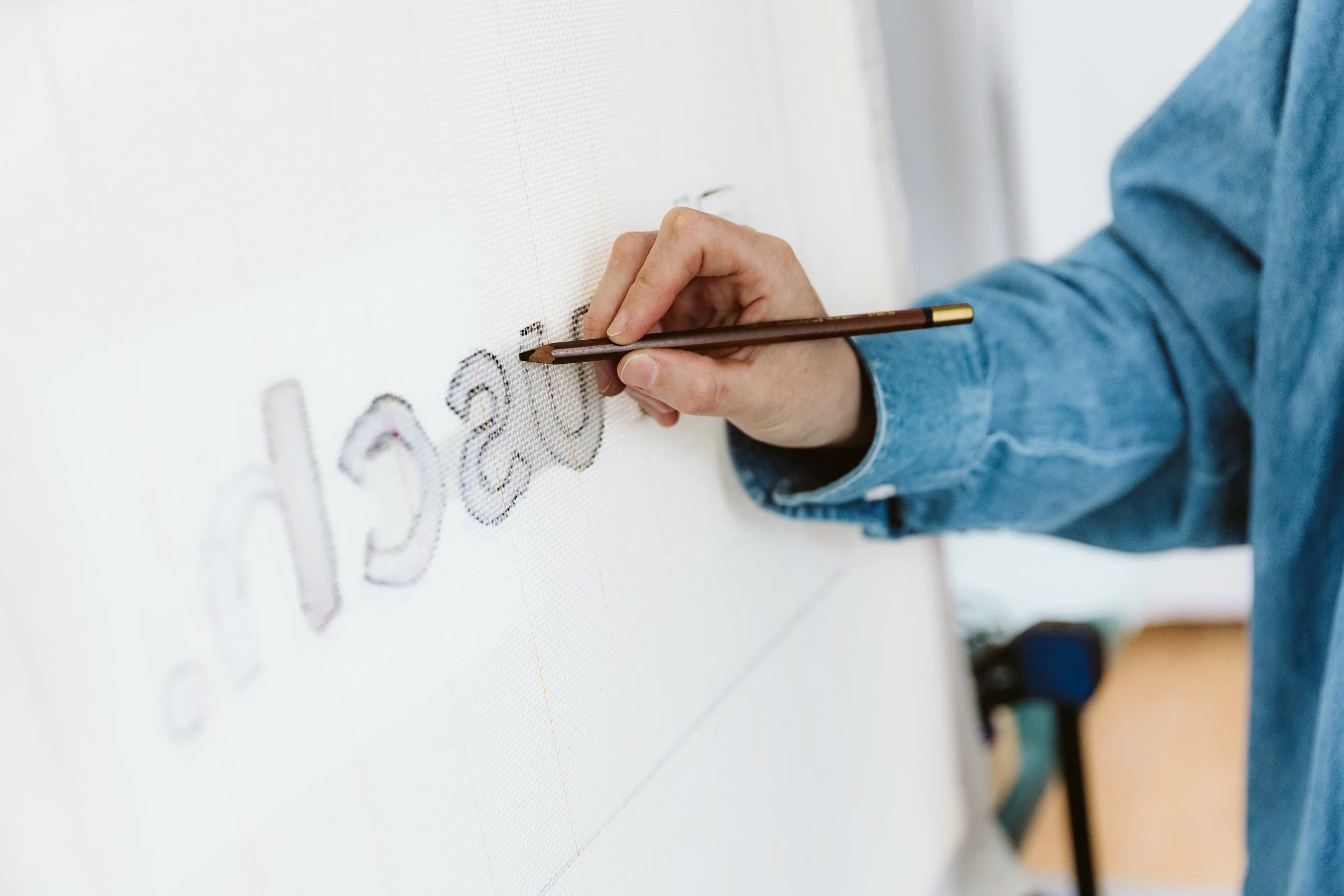 Eine Person schreibt auf ein großes weißes Blatt Papier mit einem braunen Stift. Der Fokus liegt auf der Hand und dem Stift, während die Person einen Buchstaben schreibt.