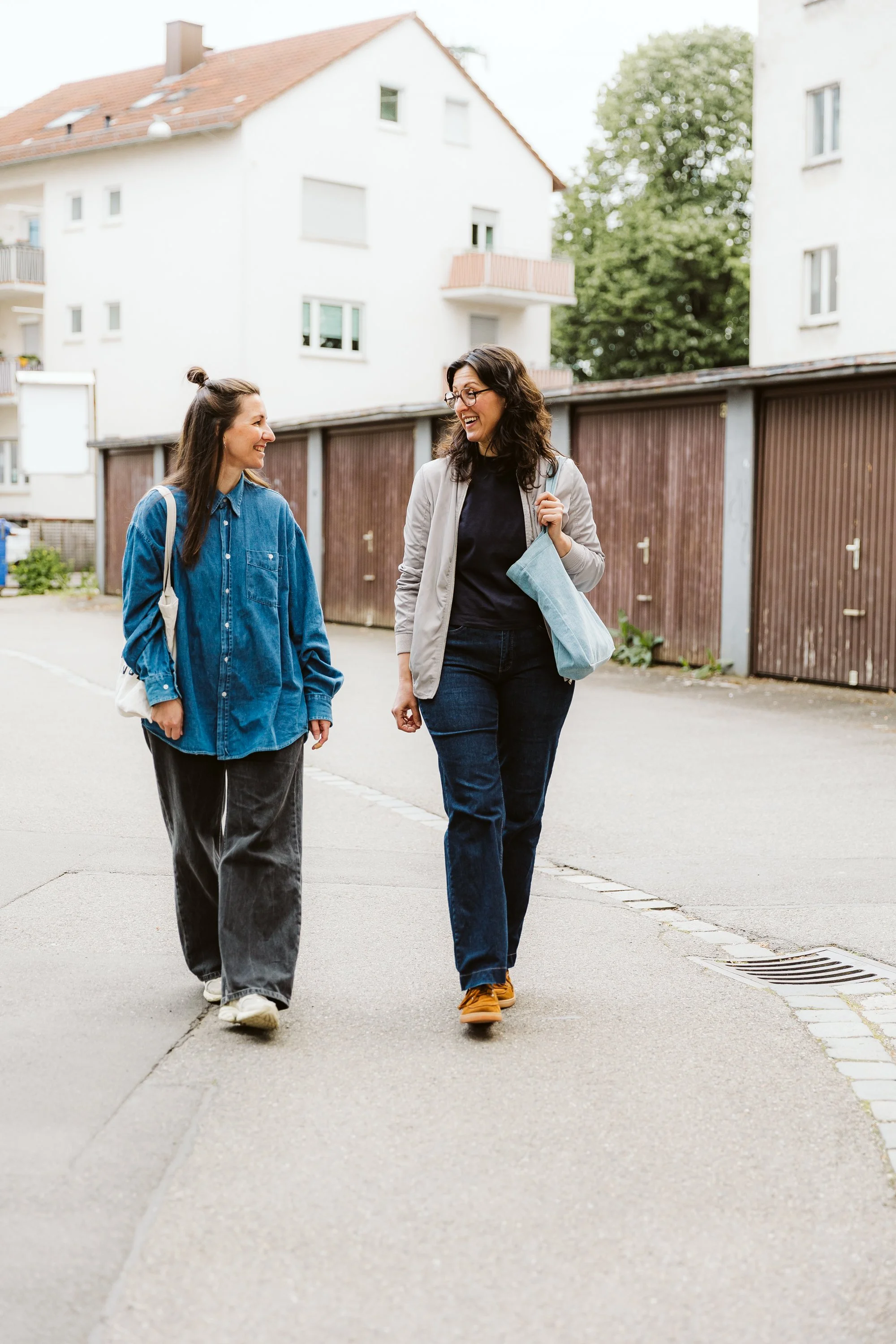 Zwei Frauen lachen und unterhalten sich beim Spazierengehen auf der Straße vor weißen Mehrfamilienhäusern und einem Baum.