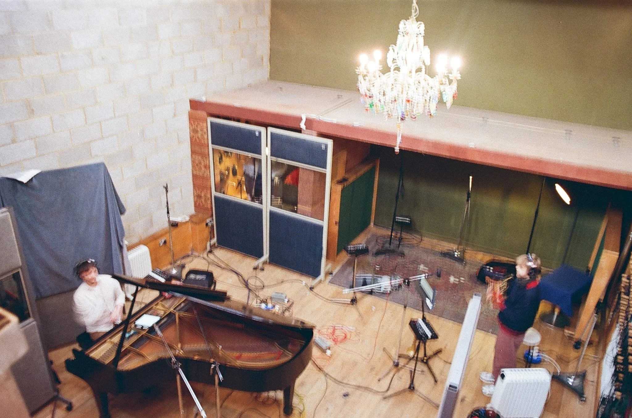 View of a recording studio with a grand piano, two people, one playing the piano and the other standing, microphones, lights, and a chandelier hanging from the ceiling.