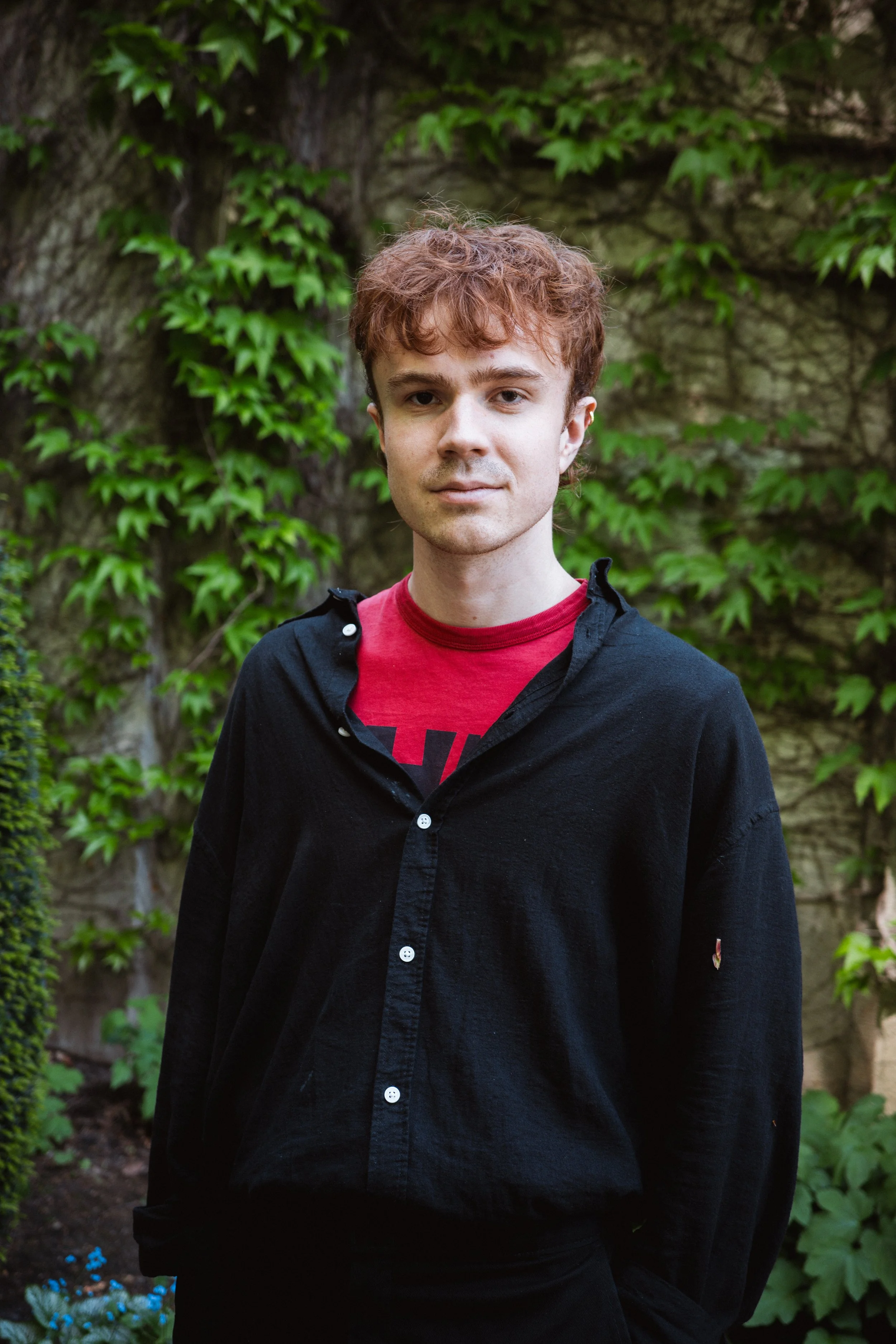 A young man with curly red hair standing outdoors in front of a stone wall covered with green ivy. He is wearing a red T-shirt and a black button-up shirt.