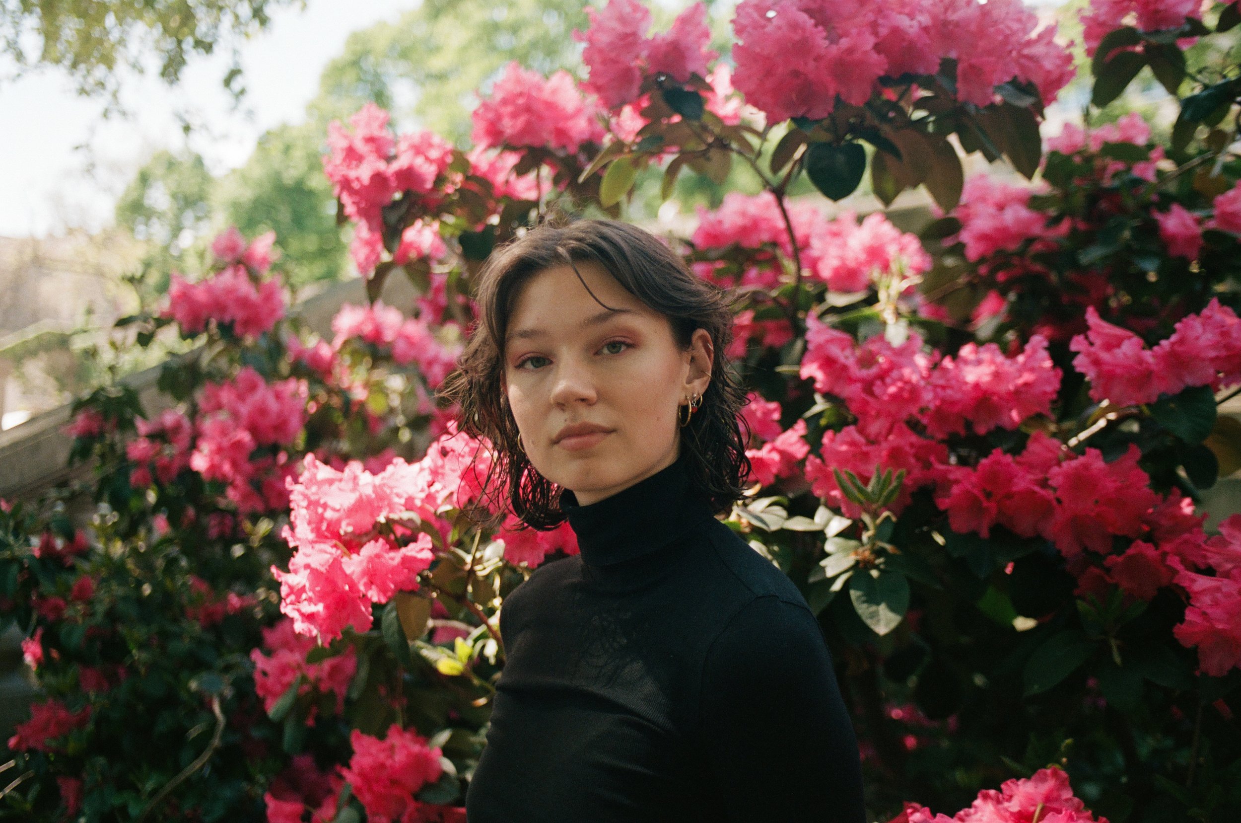 Young woman with dark wavy hair wearing a black turtleneck sweater stands in front of pink azalea flowers and green foliage.