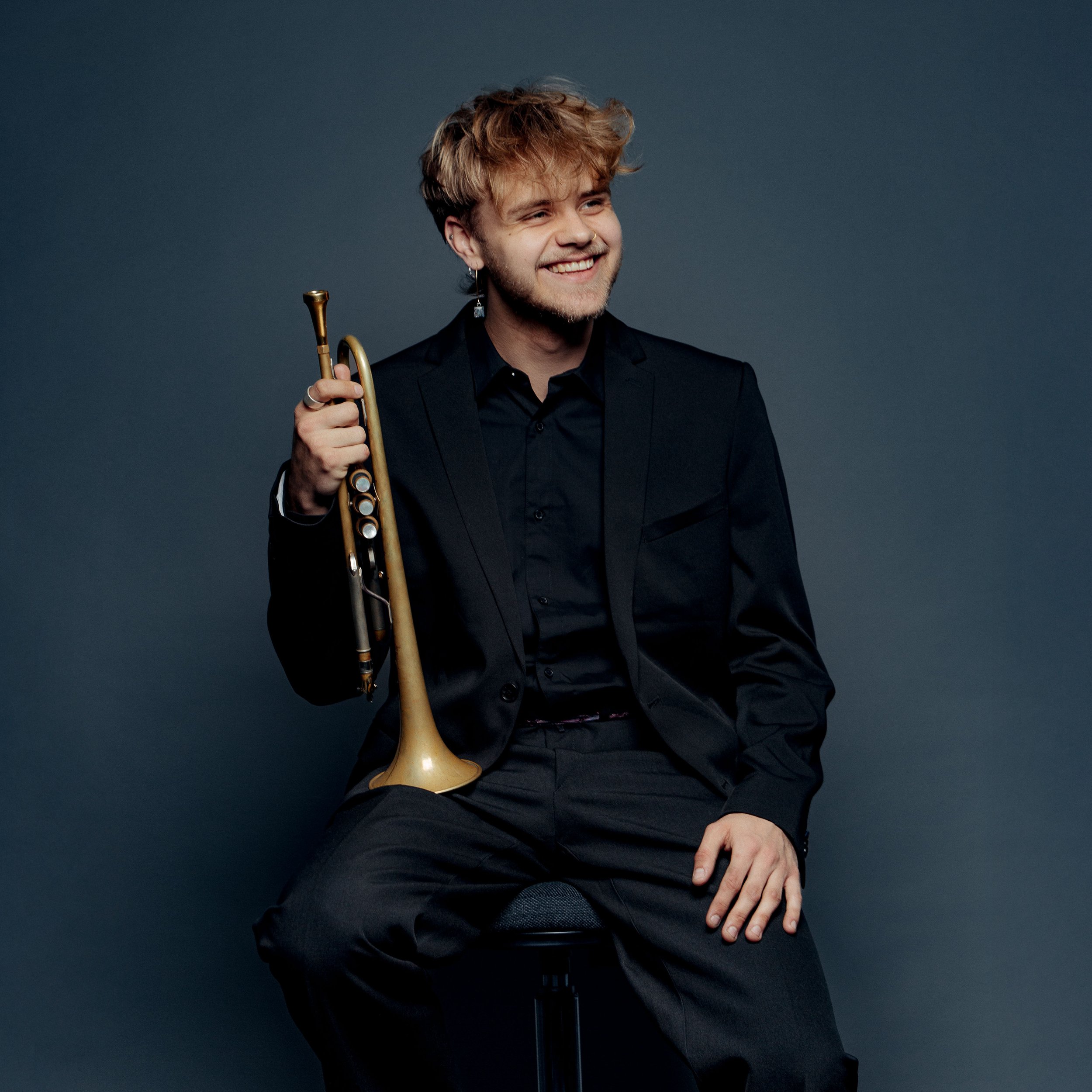 A young man with blonde, tousled hair and a beard, dressed in a black suit and shirt, smiling while sitting on a black stool against a dark background, holding a trumpet in his right hand.