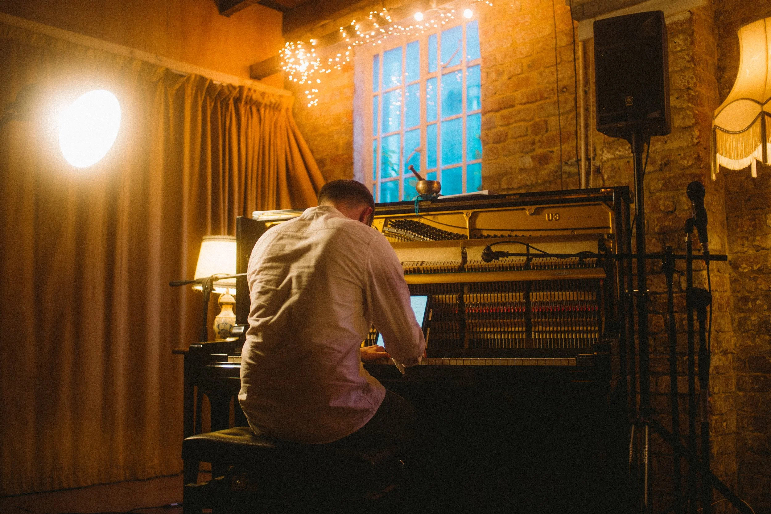 A person playing a grand piano in a warm, cozy room decorated with string lights, curtains, and a lamp, with a window and a speaker in the background.