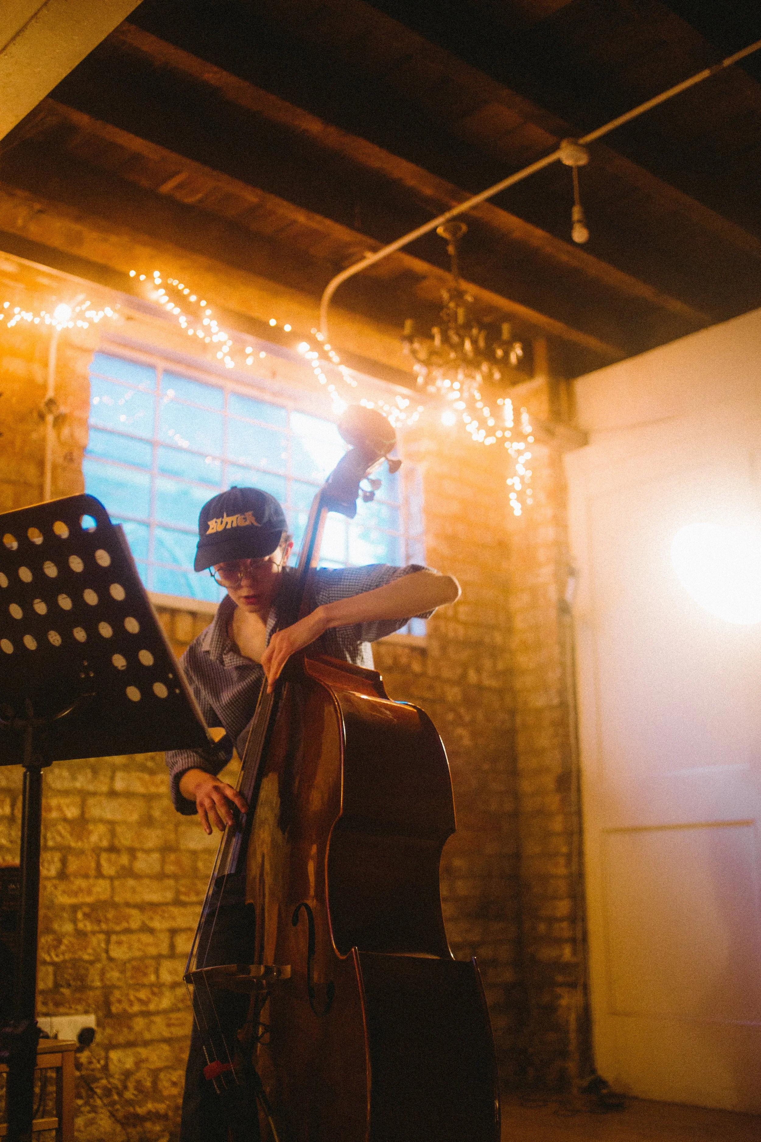 Person playing double bass in a dimly lit room with brick walls, string lights, and a window.