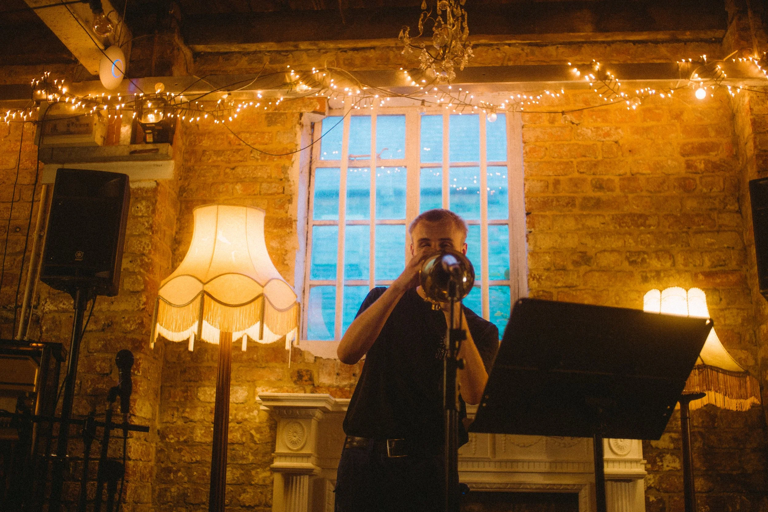 Person playing a trumpet in a warmly lit room with brick walls, vintage lamps, string lights, a window, and music stand.