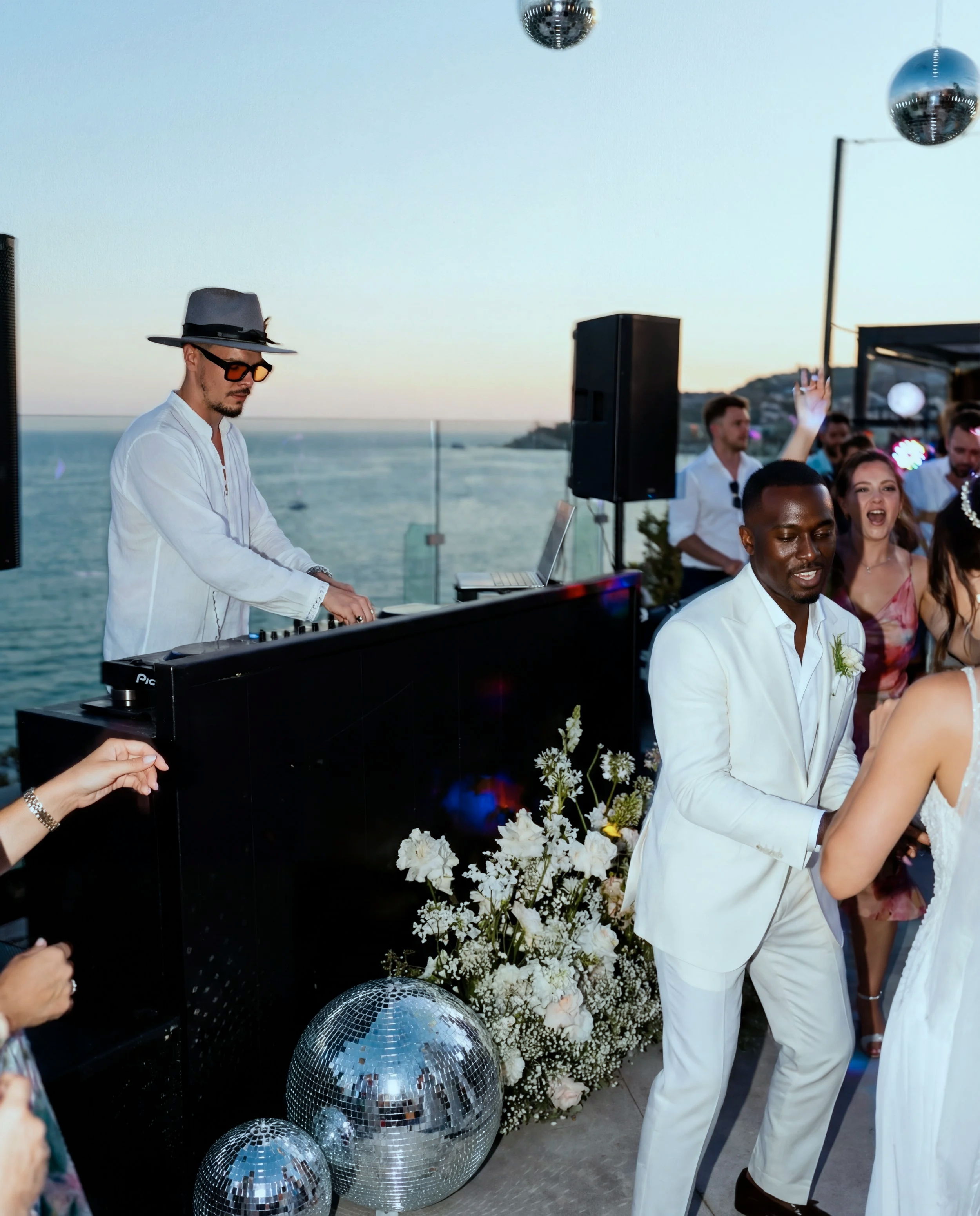 Menschen feiern eine Hochzeit am Meer mit DJ Chris PyRo in Kroatien, der Musik auflegt, und einer klassischen Hochzeitsdekoration mit weißen Blumen und Diskokugeln.