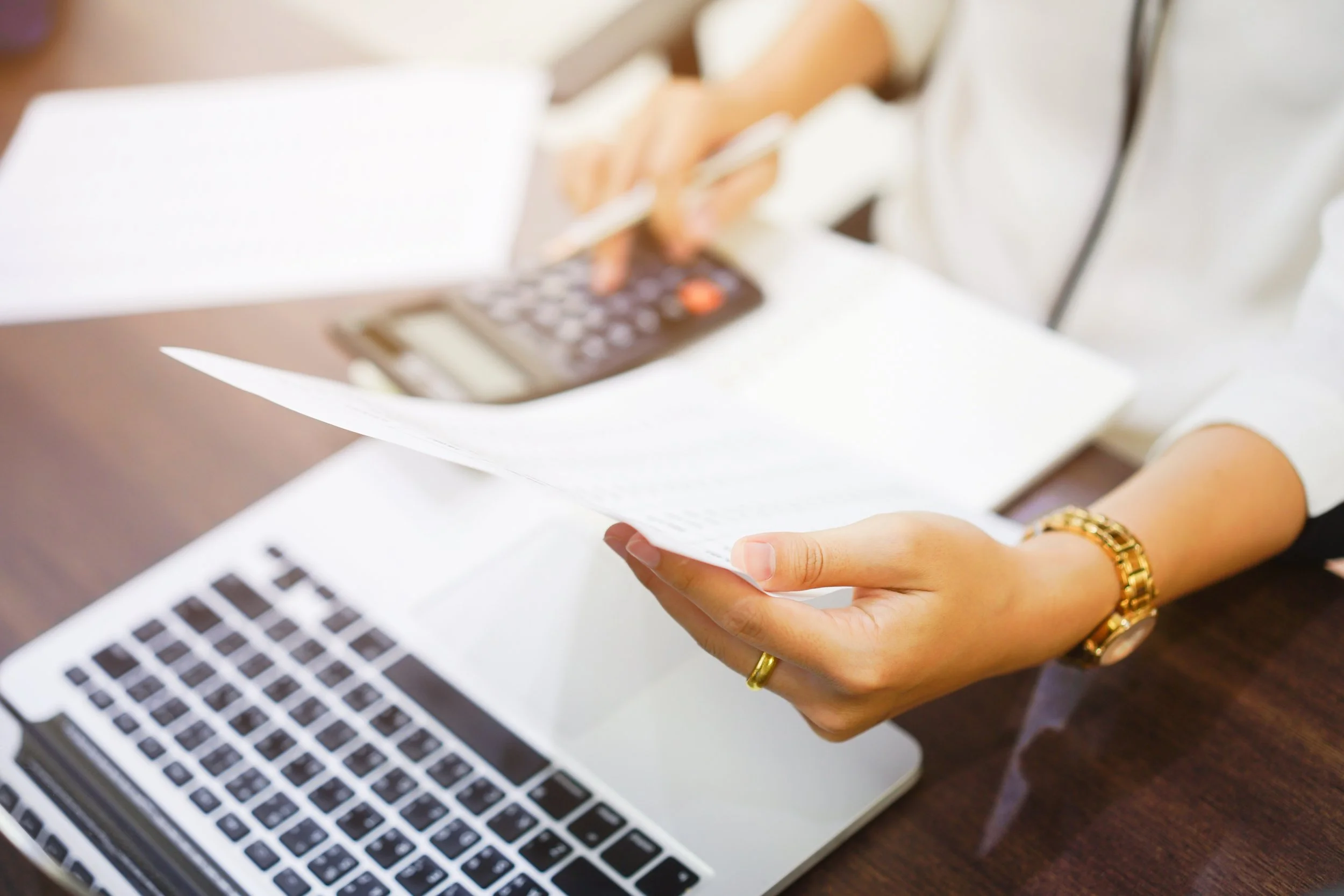 Person holding a paper with a calculator on the table, near a laptop, in an office setting.
