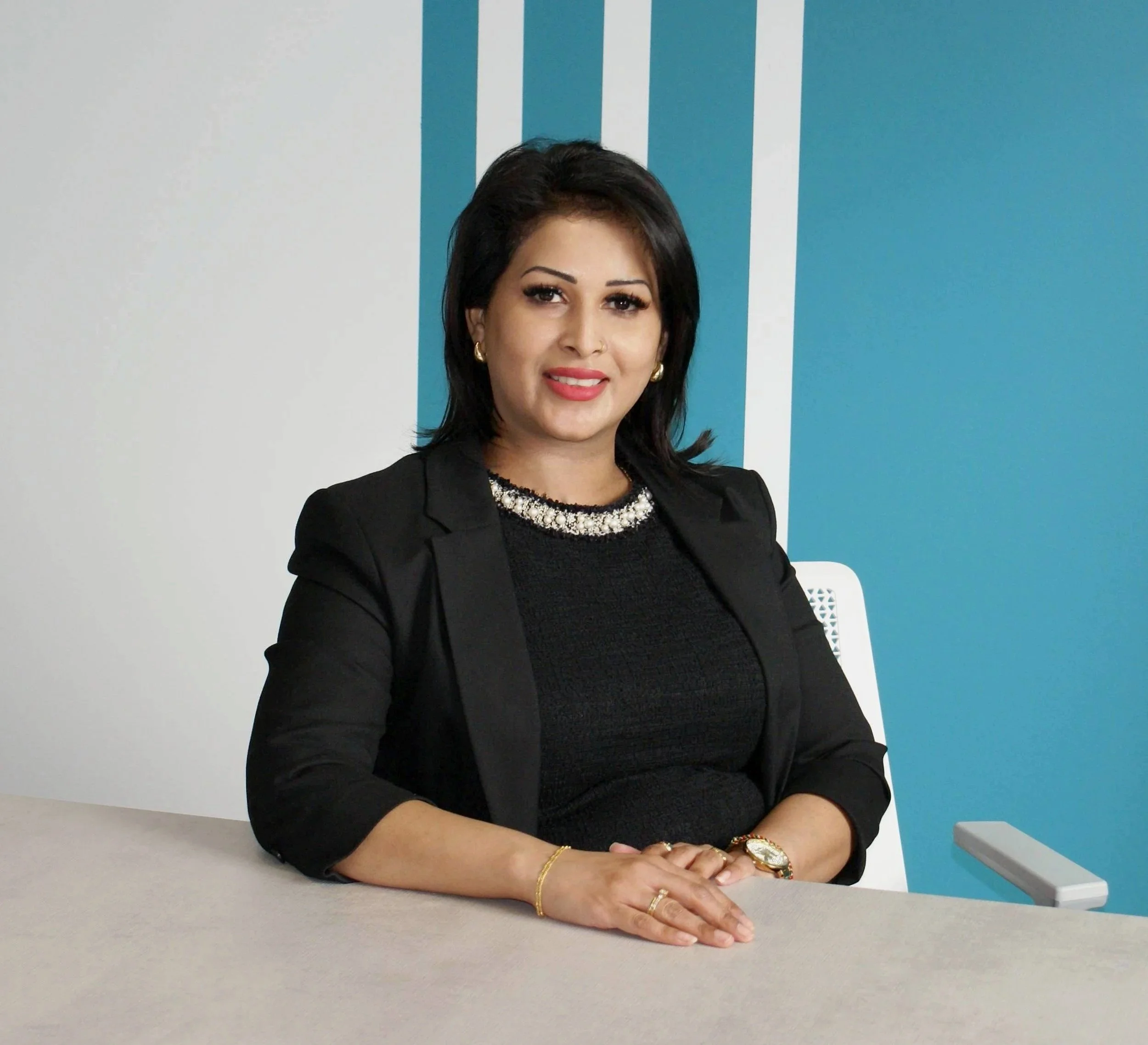 A woman sitting at a conference table, wearing a black blazer and top, with jewelry, in an office with a blue and white wall.