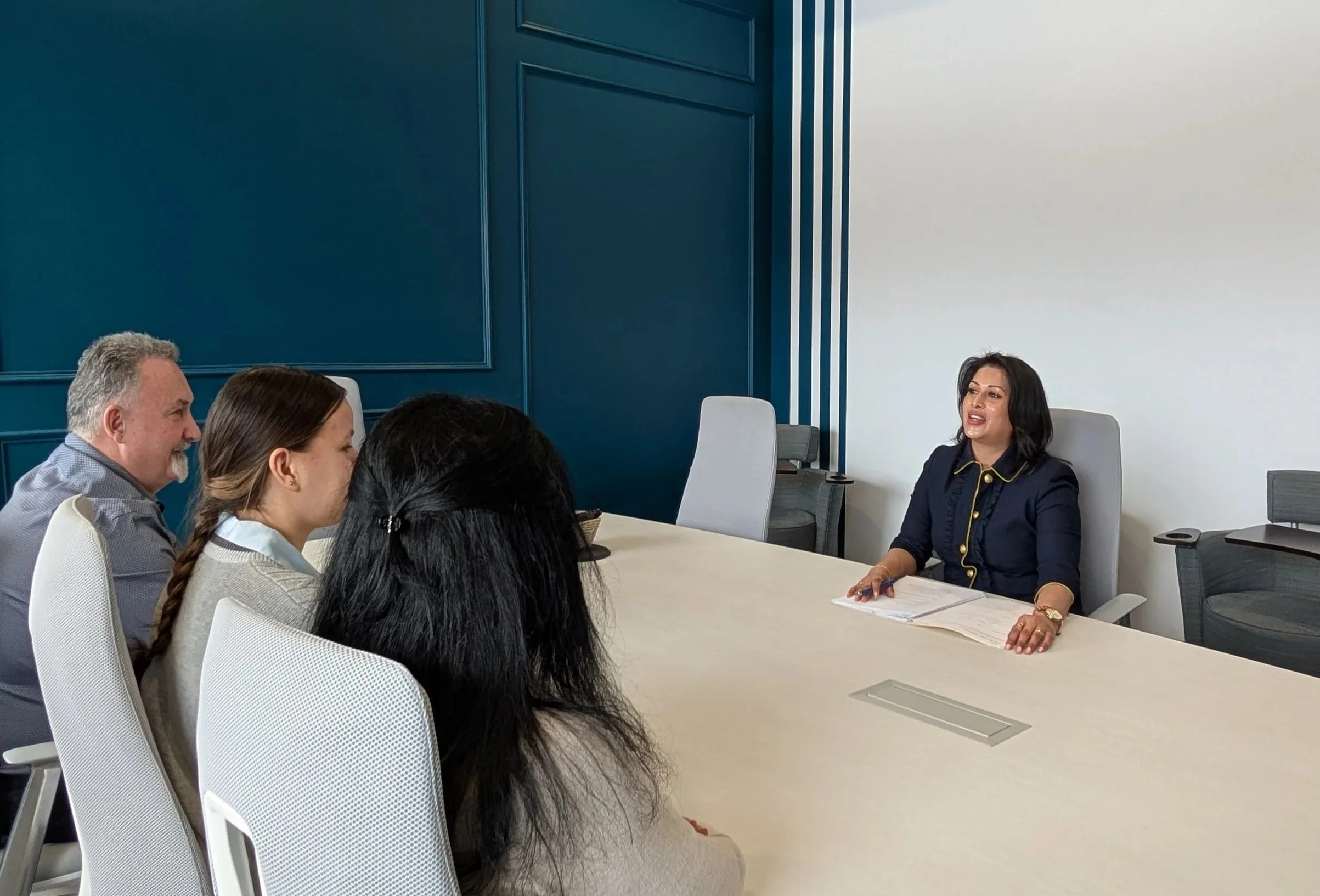A woman seated at a conference table talking to four people on the other side, with three women and one man, all listening attentively in a modern office meeting room.