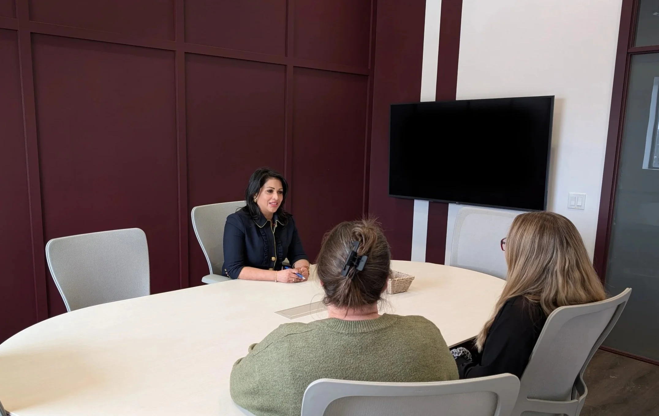 Three women having a business meeting in a conference room, one woman speaking while two others listen, with a large TV screen on the wall.