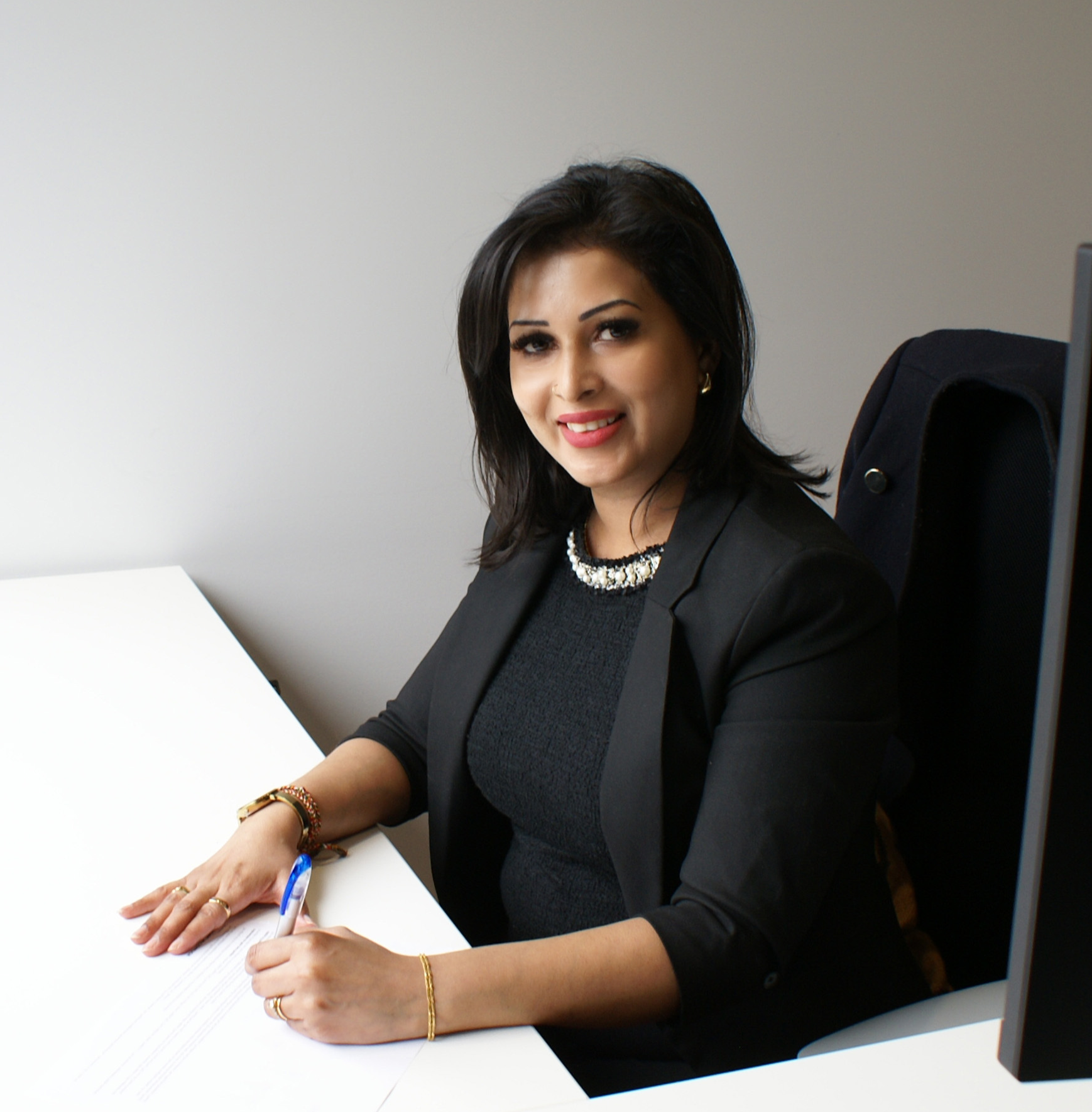 A woman with dark hair, wearing a black blazer and jewelry, sitting at a desk, signing a document with a blue pen.