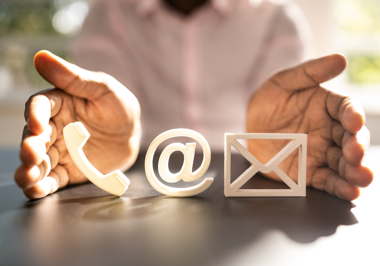 A person with dark skin reaching towards four white 3D icons representing communication methods on a table: a phone receiver, an at symbol, an envelope, and a speech bubble, in a bright room with sunlight.