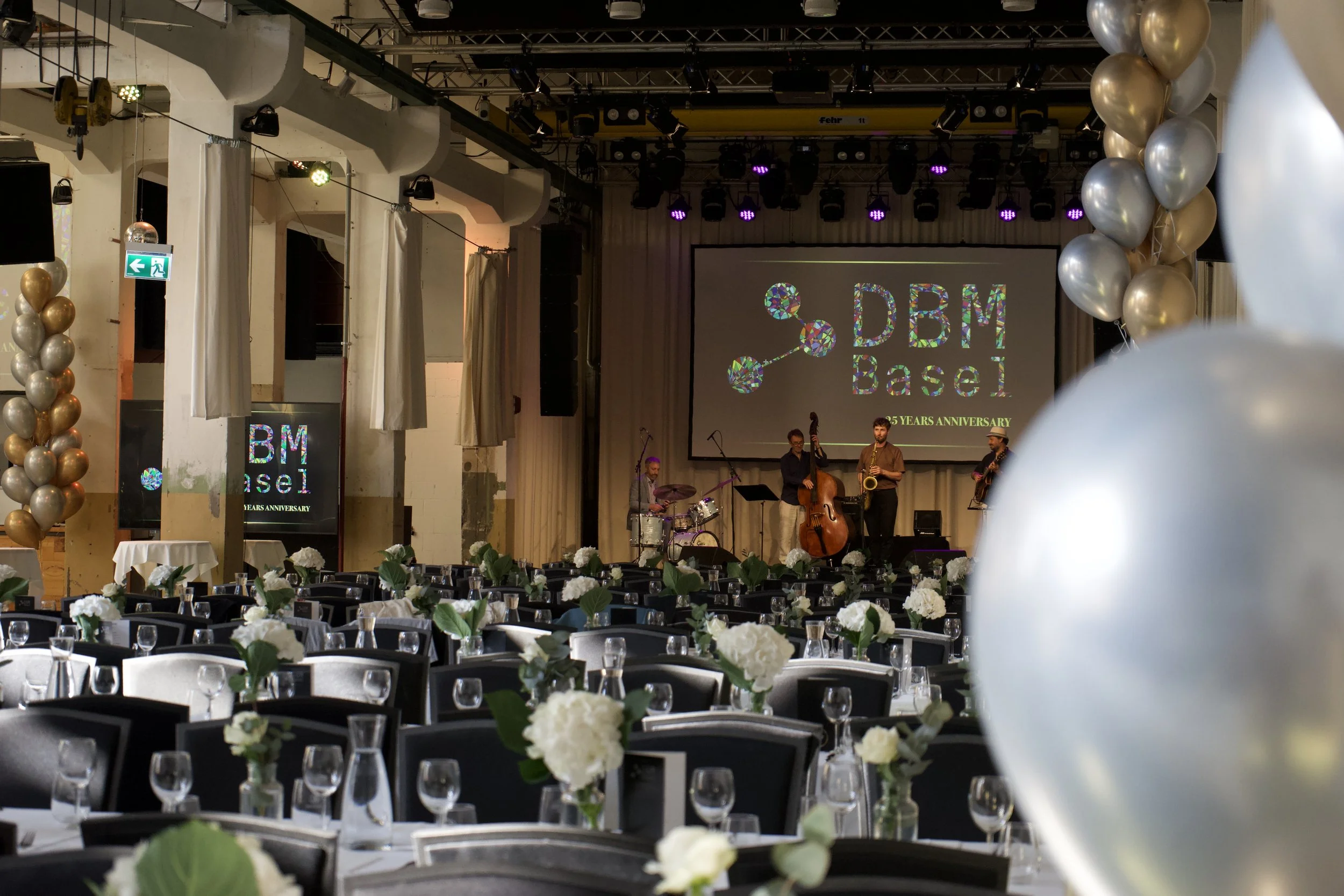 Festliche Veranstaltung im Saal mit Tischen, weißen Blumen, Ballons und einer Live-Band auf der Bühne. Projektionsschirm zeigt das Logo von DBM Basel zum 25-jährigen Jubiläum.