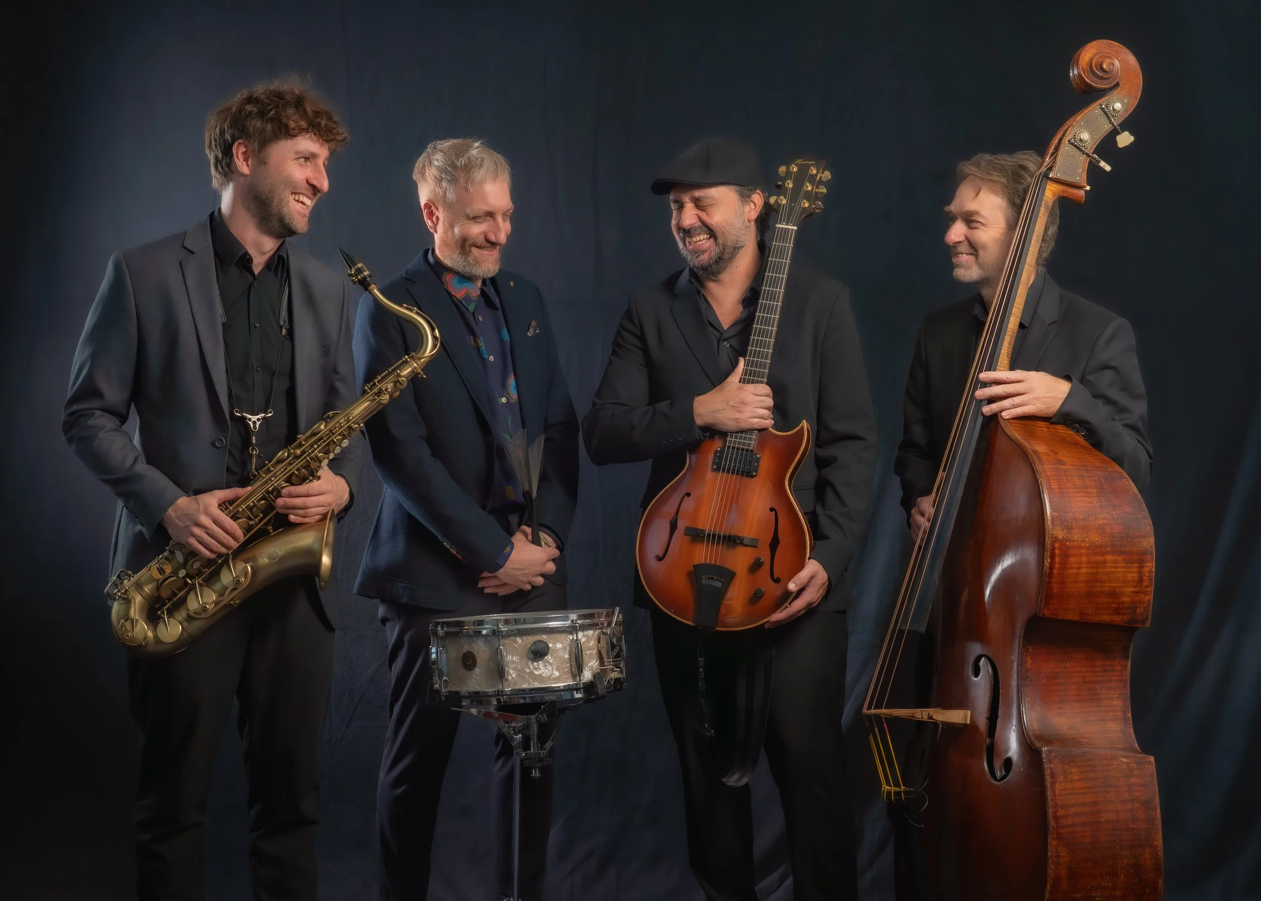 Vier Männer in dunkler Kleidung, die mit verschiedenen Musikinstrumenten lachen und zusammenstehen. Eines hält Saxophon, ein anderes eine Trommel, ein drittes eine E-Gitarre und das letzte ein Kontrabass.