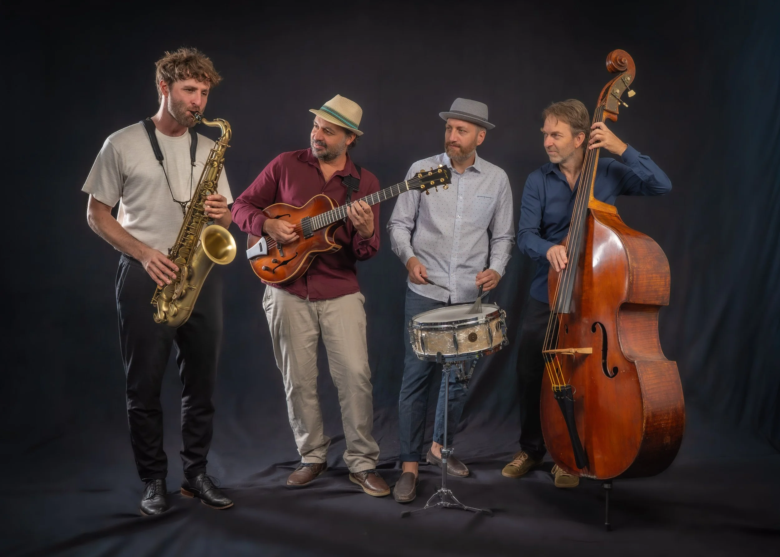 Vier Musiker mit verschiedenen Instrumenten vor dunklem Hintergrund, darunter Saxophon, Gitarre, Tamburin und Kontrabass.
