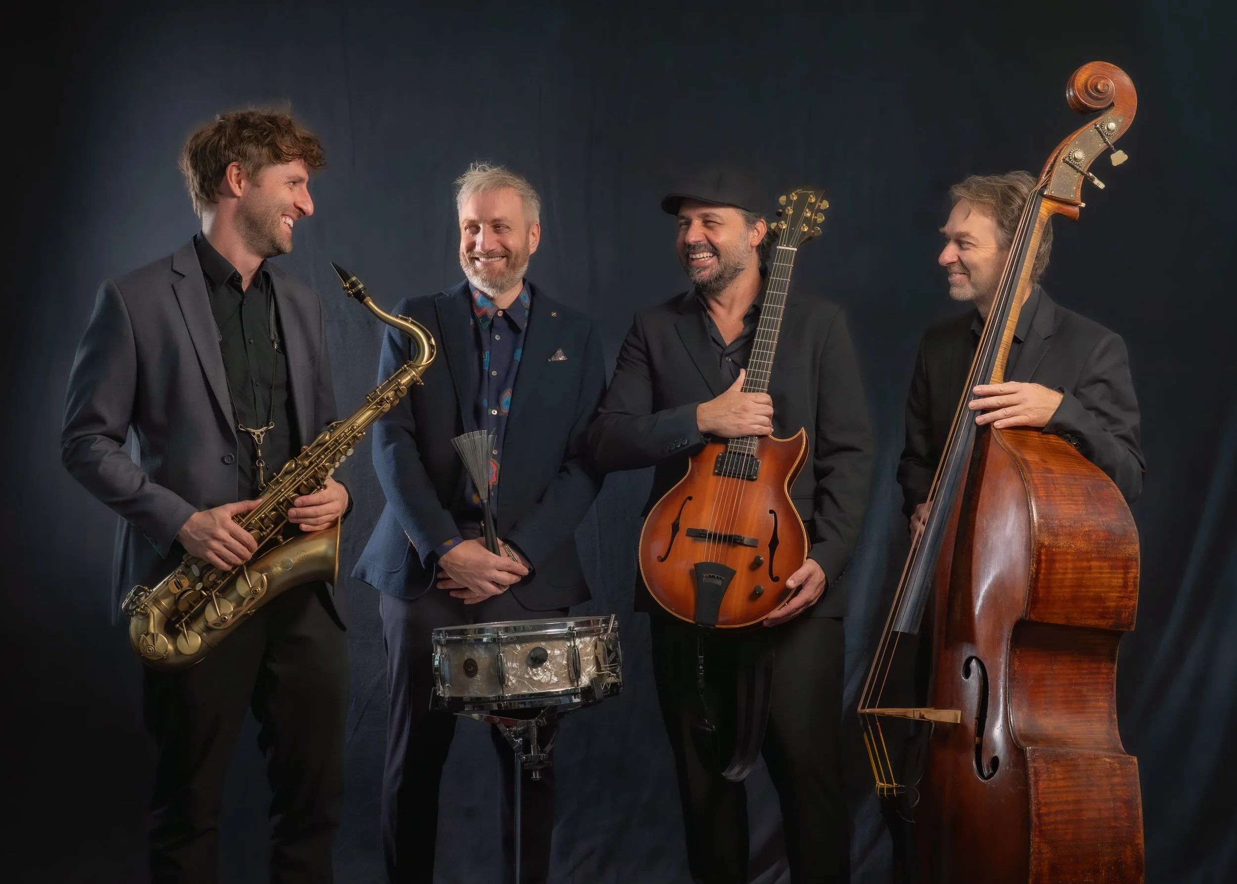 Vier Männer mit Musikinstrumenten, lachen und unterhalten sich vor dunklem Hintergrund.