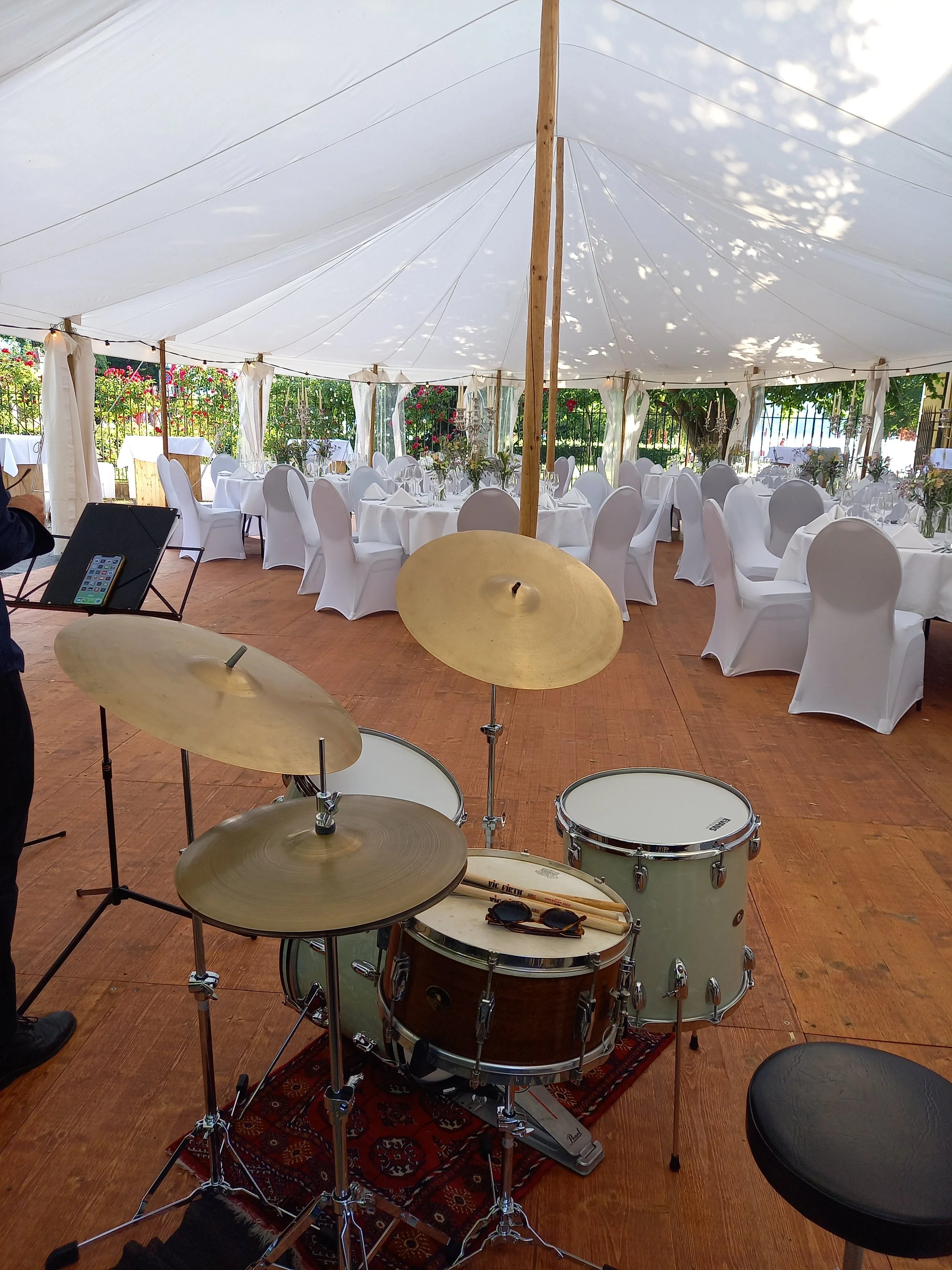 Ein Zelt mit weißen Tischtüchern, weißen Stühlen und Blumen auf den Tischen, Blick auf die leere Tanzfläche und einen Schlagzeug im Vordergrund.
