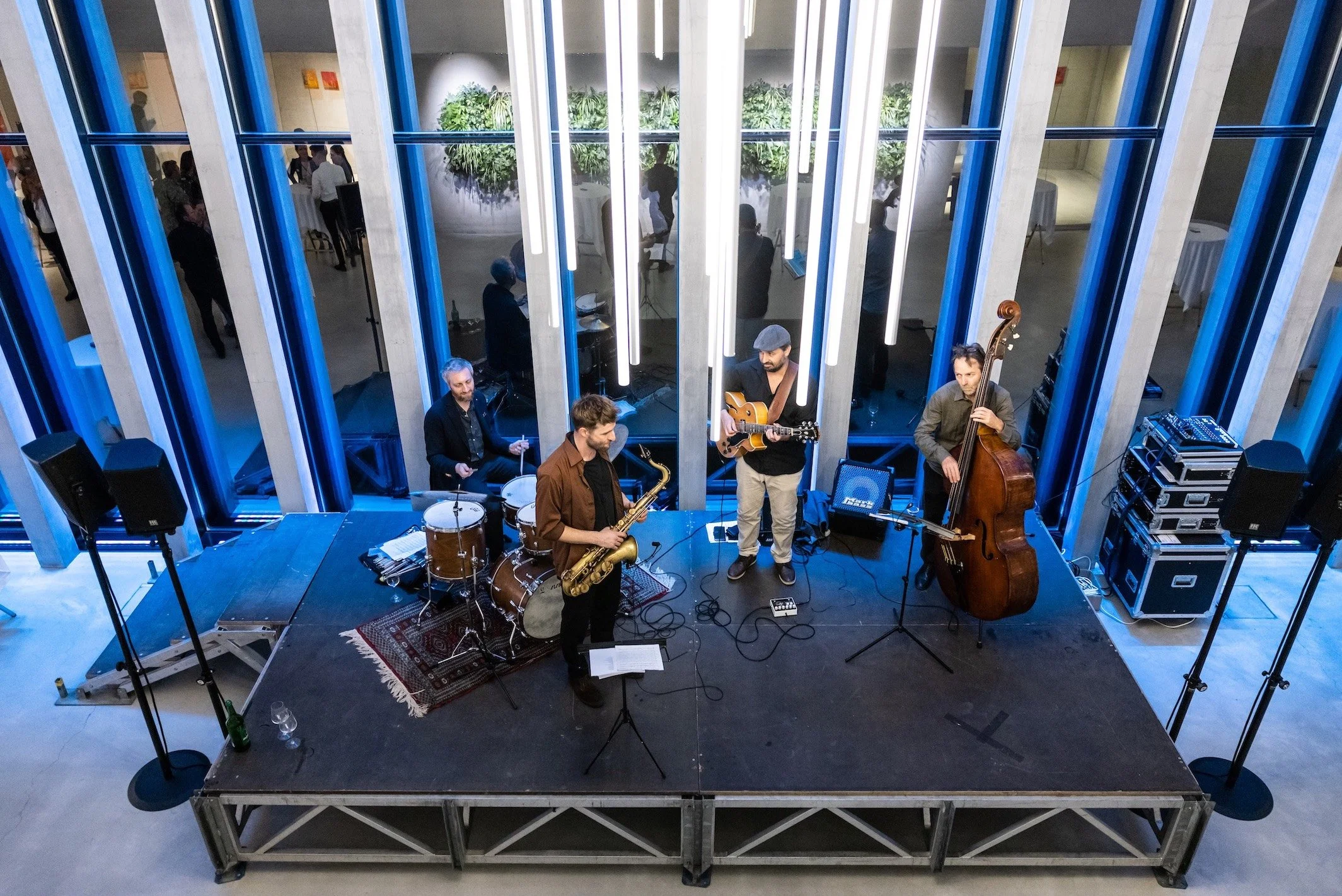 Jazzband mit fünf Musikern auf einer Bühne bei einer Veranstaltung, im Hintergrund Gäste und Pflanzen sichtbar.