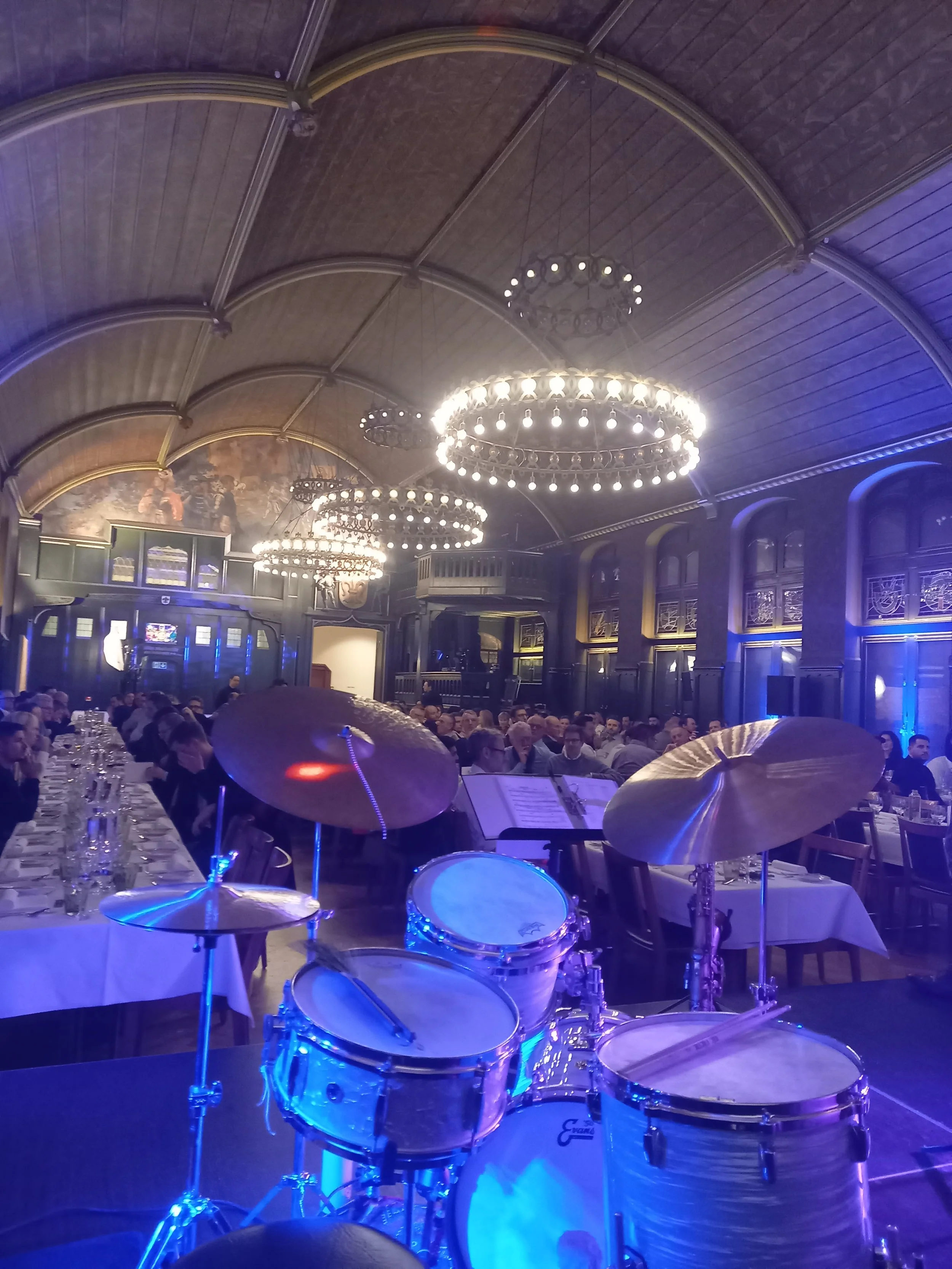 Ein Saal mit Tischen und vielen Gästen, im Vordergrund eine Trommel und zwei Becken, Bühnenmusik-Equipment, hohe Decke mit Kronleuchtern, dunkle Wände mit Fenstern und Wanddekorationen, bläuliches Licht.