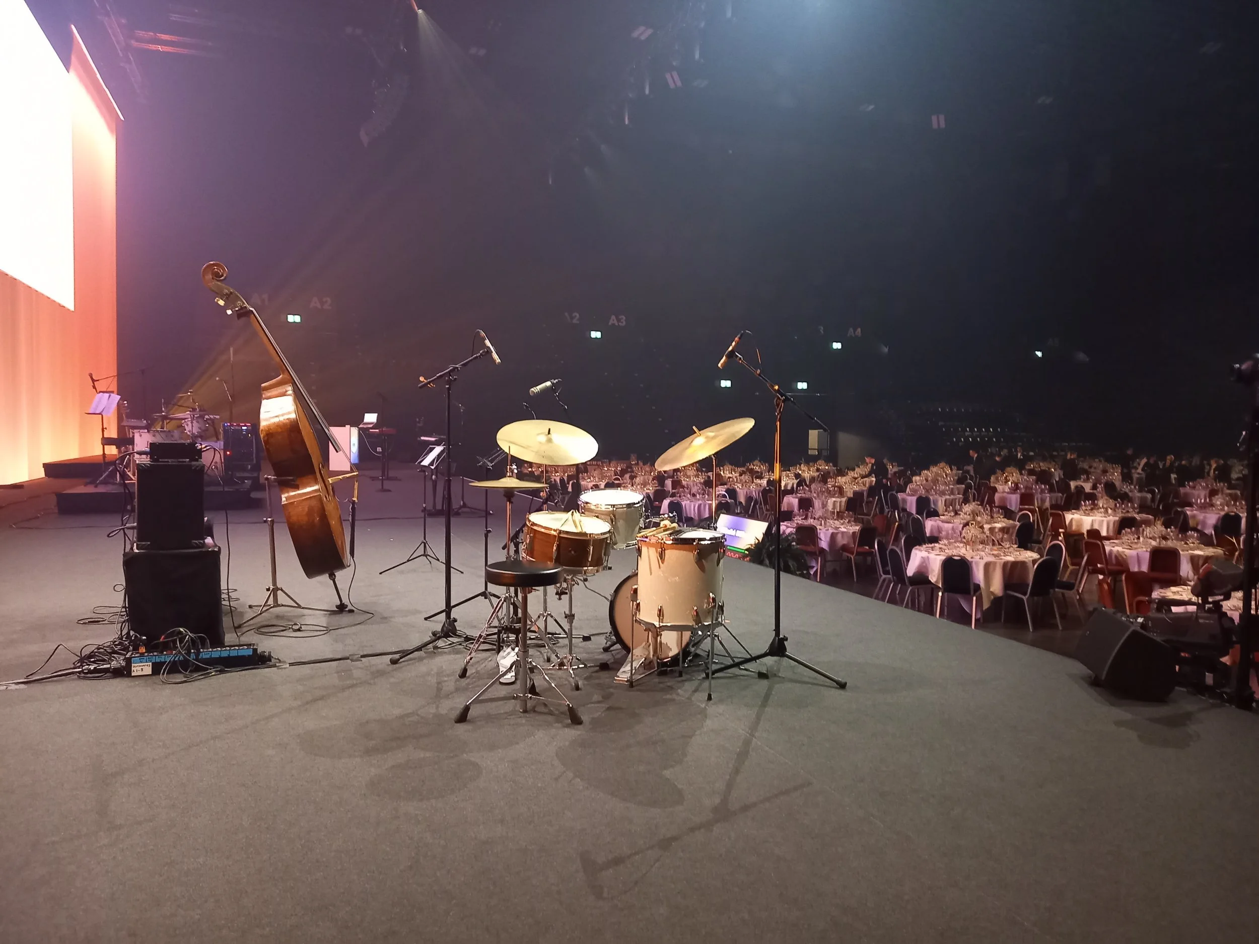 Auf einer Bühne sind Musikinstrumente wie Schlagzeug, Kontrabass und Mikrofone aufgebaut, vor einem großen Saal mit zahlreiche gedeckten Tischen.