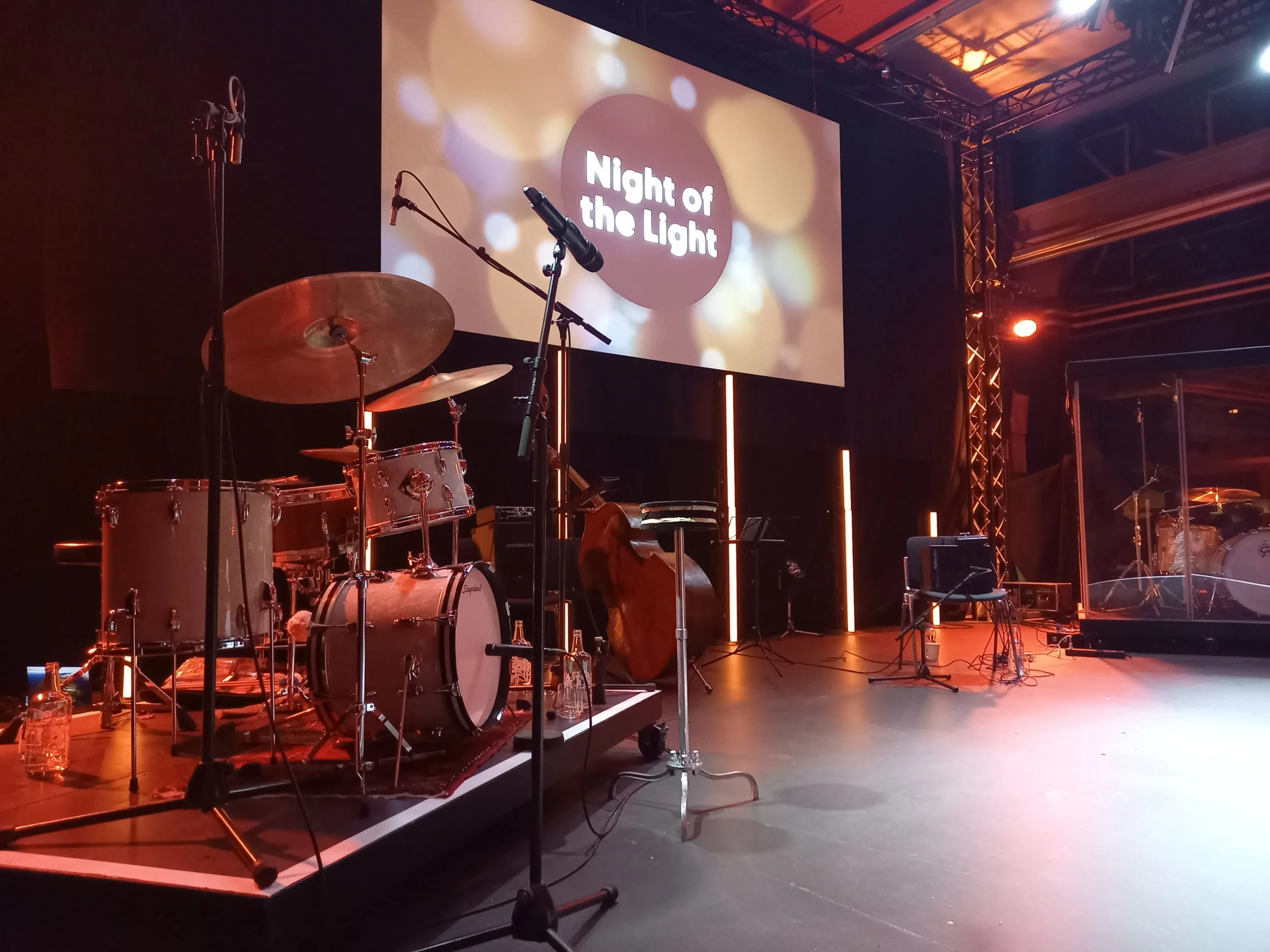 Bühne mit Musikinstrumenten, darunter Schlagzeug und Kontrabass, vorbereitet für eine Veranstaltung mit dem Titel 'Night of the Light', auf einem großen Bildschirm im Hintergrund.