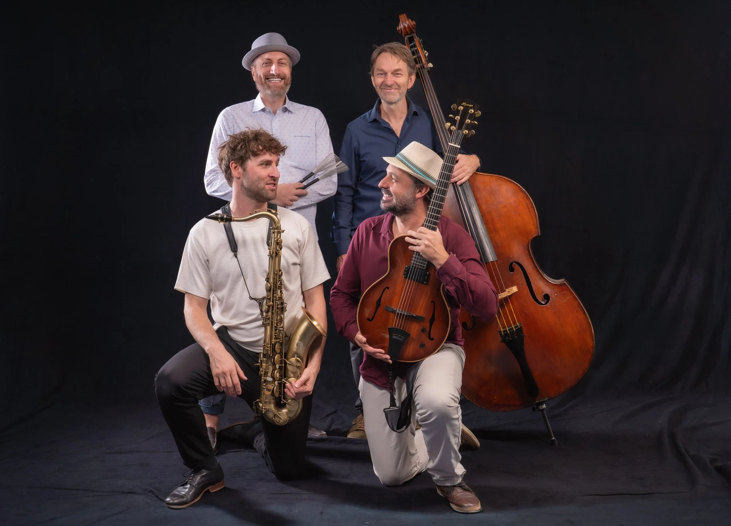 Fünf Männer mit Musikinstrumenten, einige stehen, andere knien, vor schwarzem Hintergrund, lachend und in lockerer Kleidung, mit einem Kontrabass, Saxophon, Gitarre und einer kleinen Trommel.