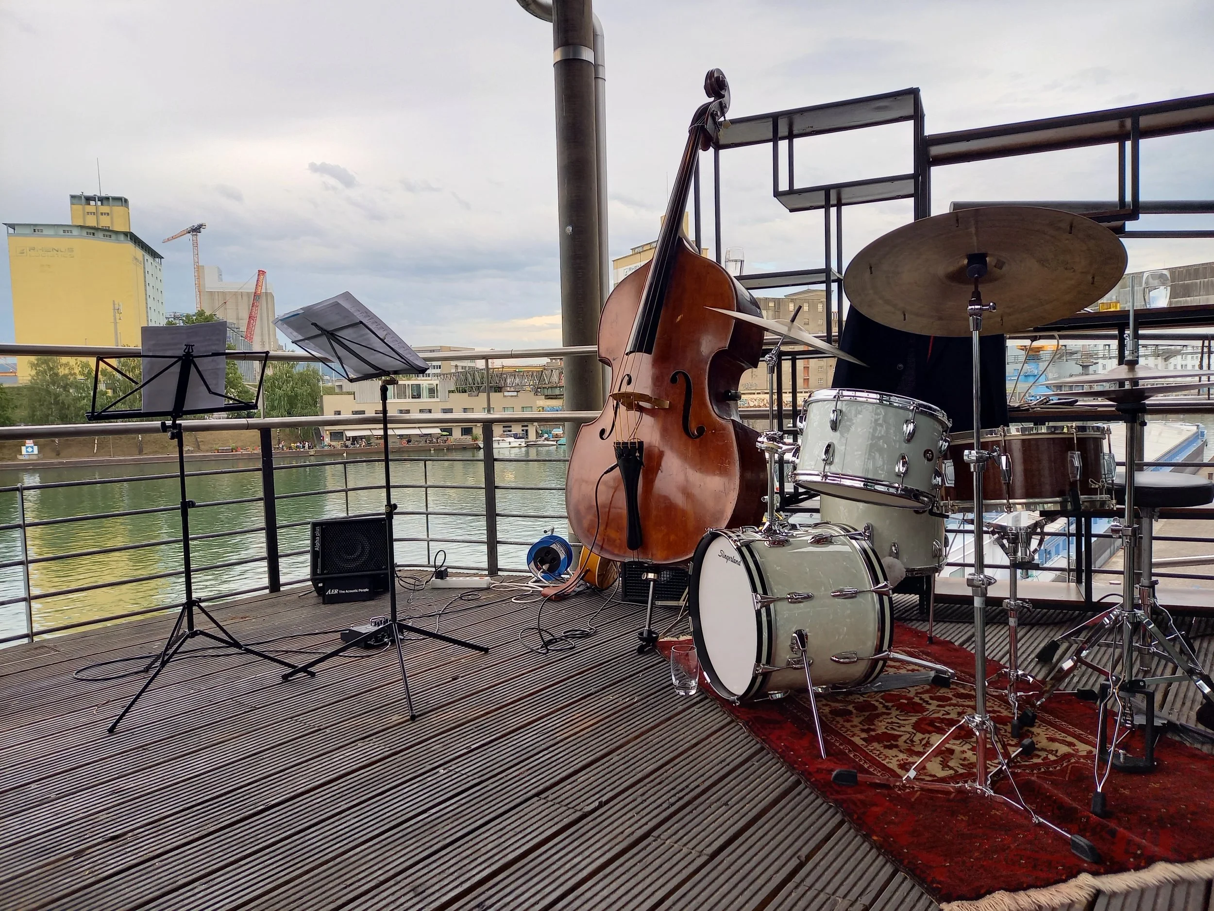 Musikerinstrumente auf einer Terrasse am Wasser, einschließlich Schlagzeug, Kontrabass, Notenständer, Mikrofon und einem kleinen Verstärker, vorbereitet für eine Aufführung.