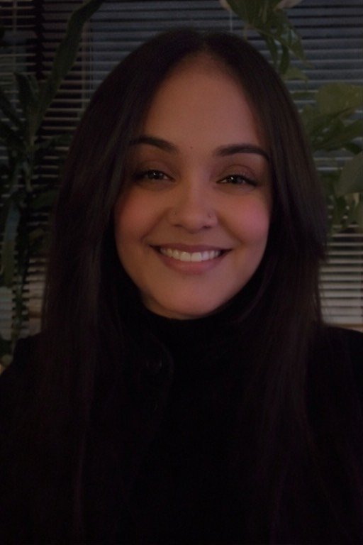 A young woman with long dark hair smiling in front of a window with blinds and green plants.