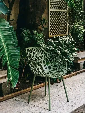 A modern outdoor chair with an intricate lattice backrest and armrest, placed on a patio next to lush green plants and a tree.