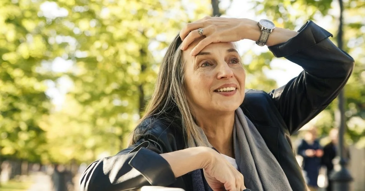 older woman holding her hand to her head and smiling. Background of trees