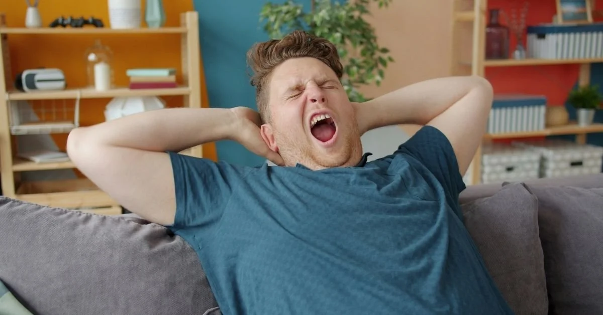 man yawning and stretching with blue top sitting on sofa