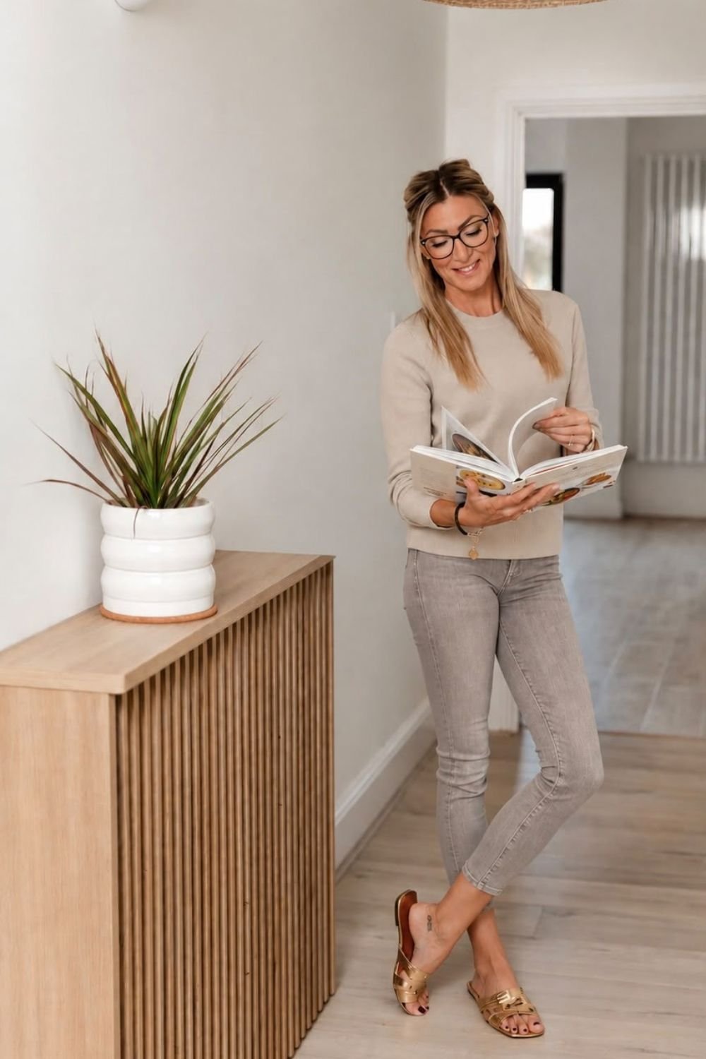 woman wearing cream jumper and faded grey jeans, holding and reading a book.