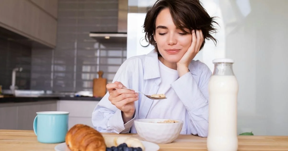 woman with dark hair smiles and looks at spoon as she eats cereal from a bowl. Milk, pastries and fruit on the table in front of her