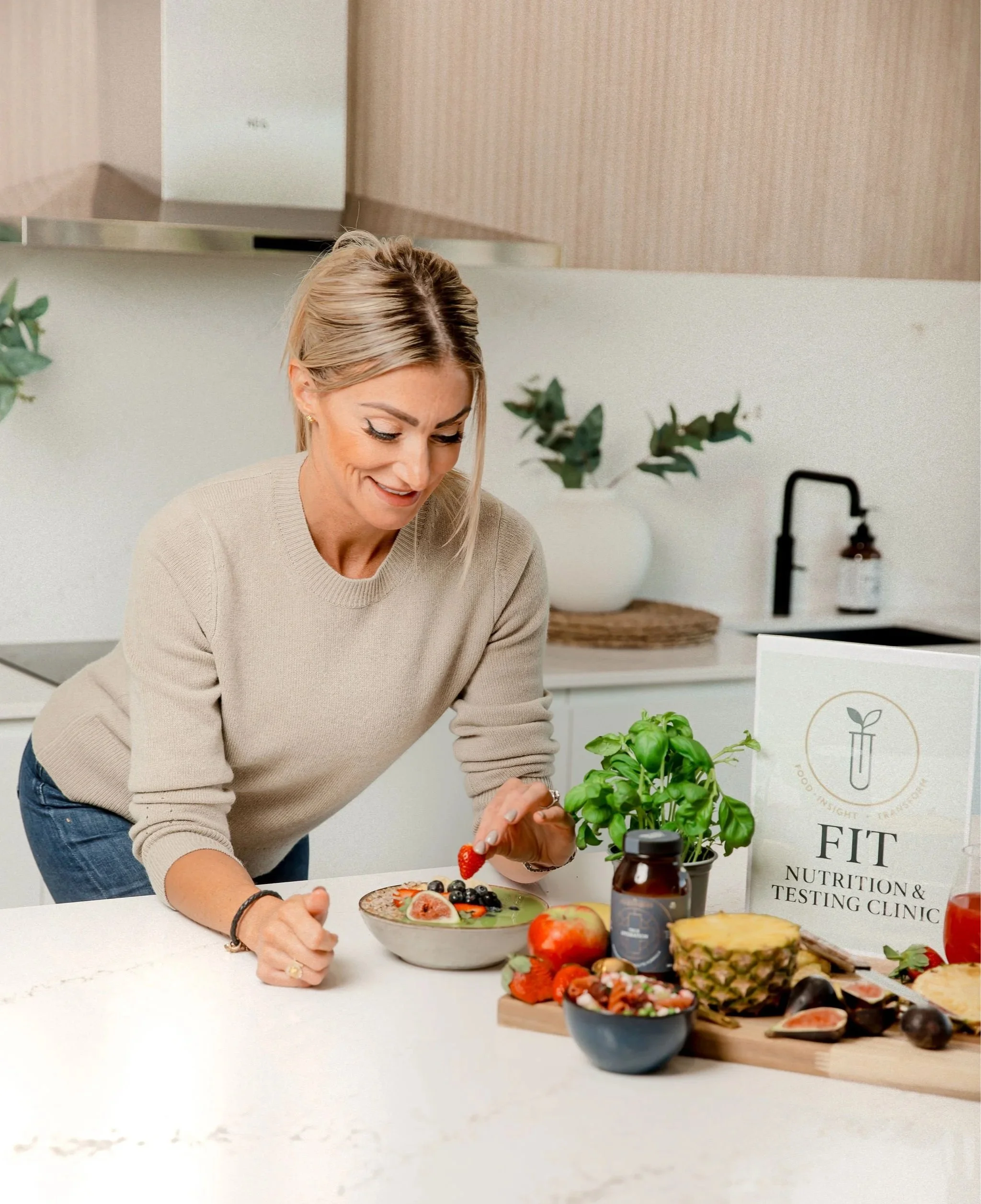 Becki Hawkins in a kitchen, placing strawberry into a healthy delicious bowl of food, smiling and looking down at her creation.