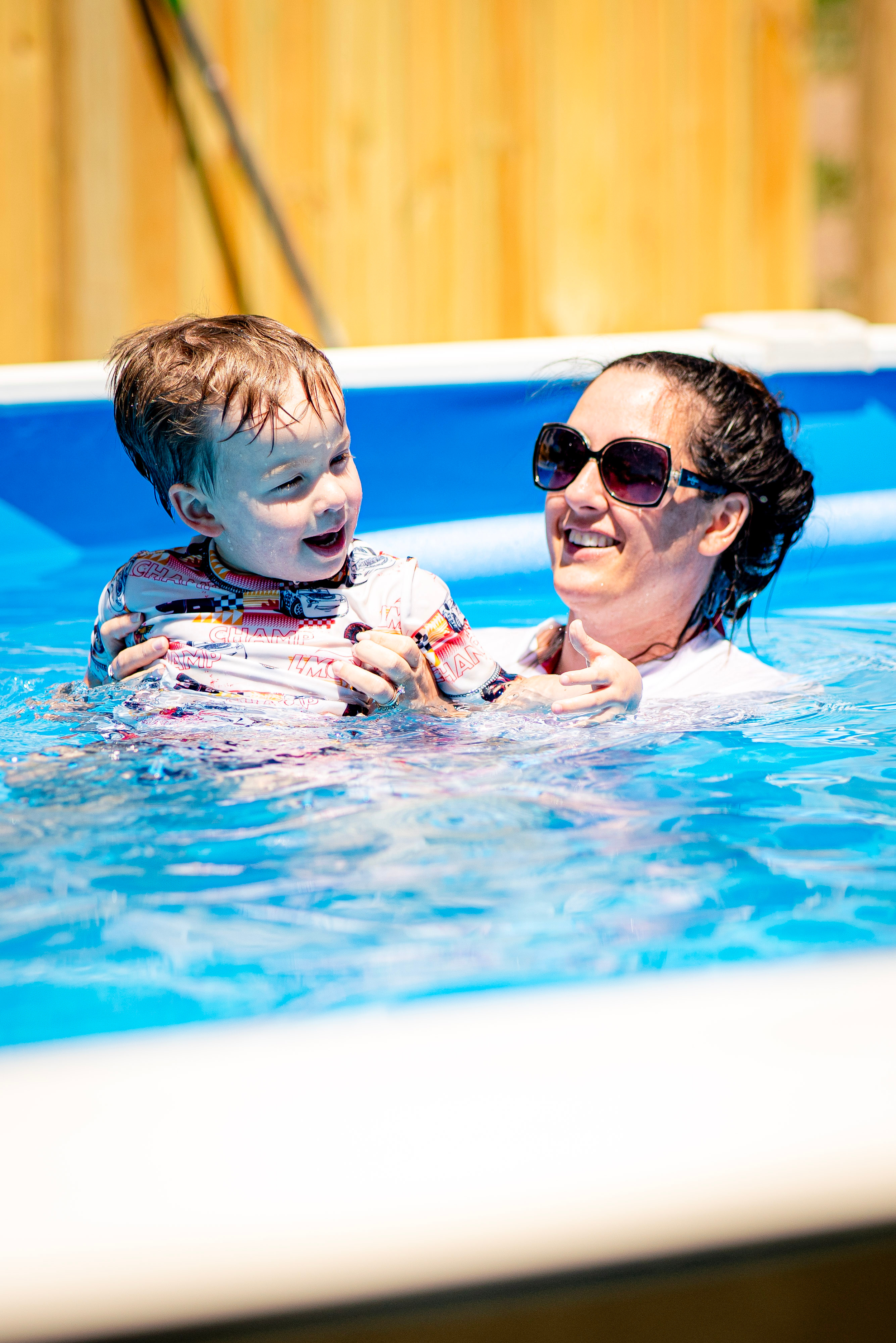 A woman with dark hair and sunglasses smiling while holding a young boy with wet hair in a swimming pool, both enjoying swimming outdoors.
