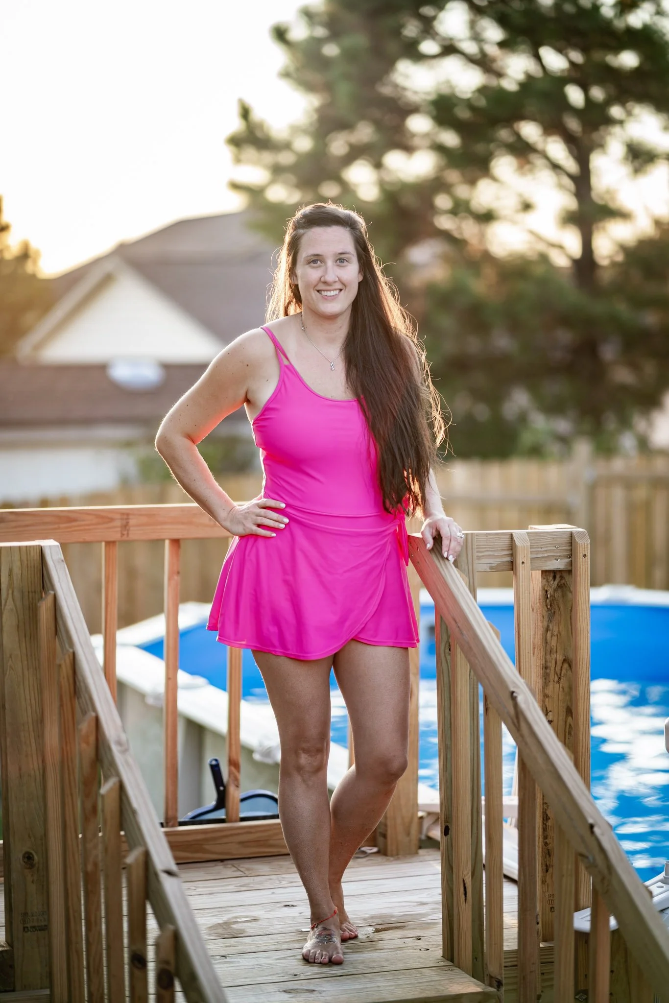 A woman in a bright pink dress standing on a wooden deck by a swimming pool during the late afternoon or early evening.