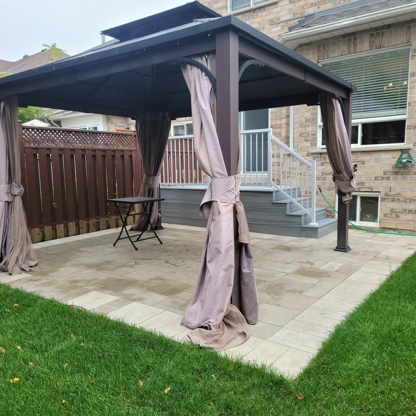 Backyard patio with a dark brown gazebo with curtains, a small folding table, pavers, a wooden deck with stairs, a brick house wall, and a grassy lawn.