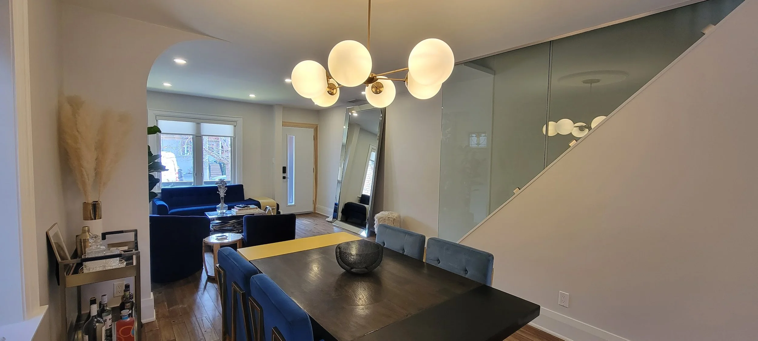 Modern dining area with dark wood table, blue upholstered chairs, a black wire bowl centerpiece, and a large angular mirror leaning against the wall. A gold and glass globe chandelier hangs above the table. In the background, a sitting area with a blue velvet sofa, a window, and a front door.