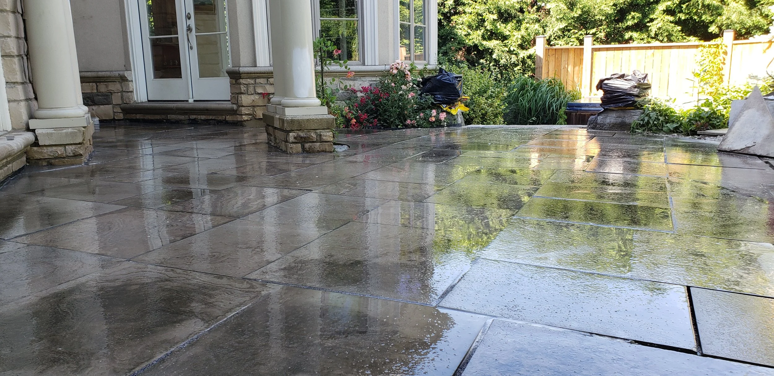 Wet stone patio in front of a house with flower beds and a wooden fence in the background.