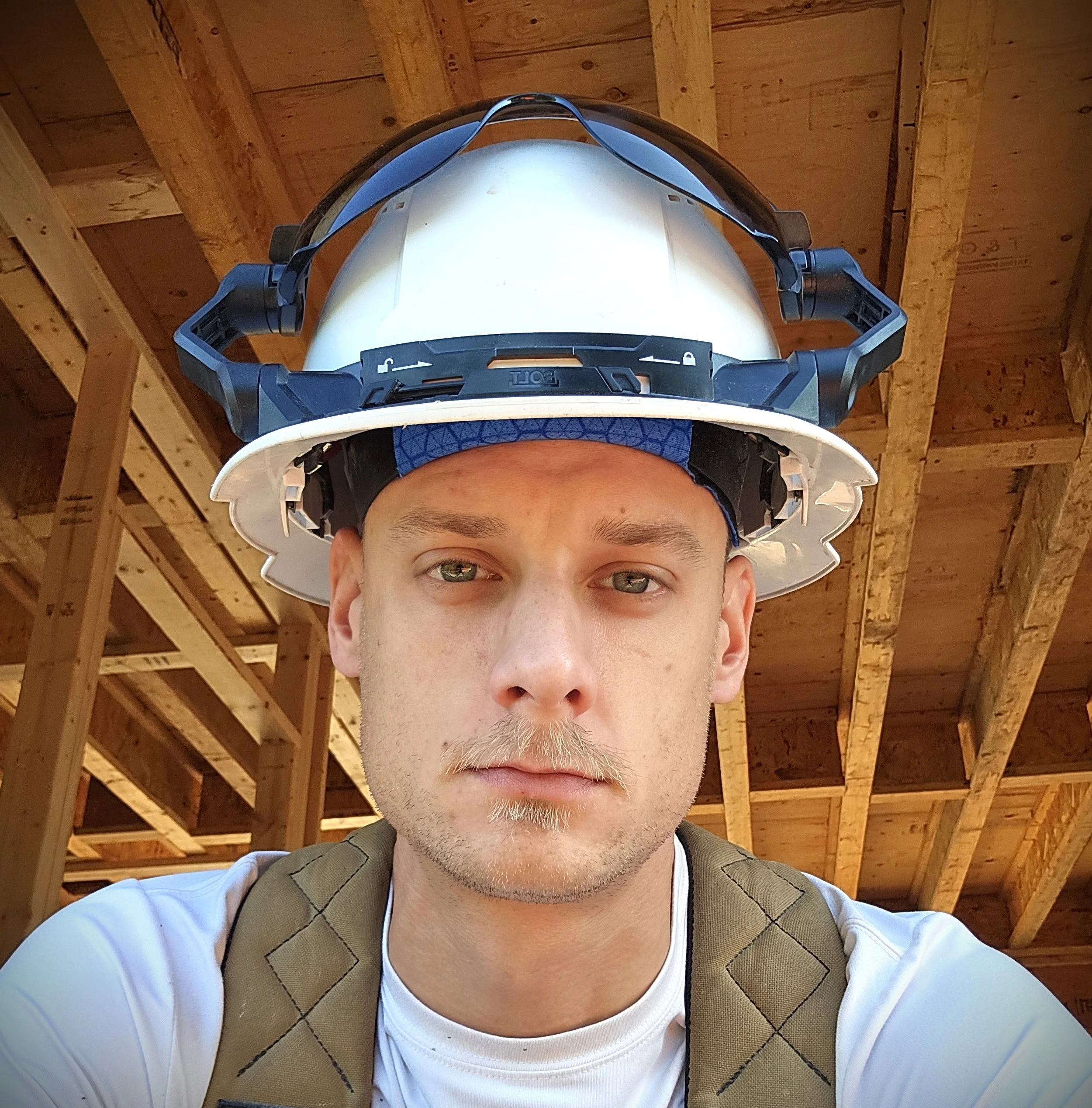 A man wearing a white hard hat and safety goggles on a construction site with wooden framing in the background.