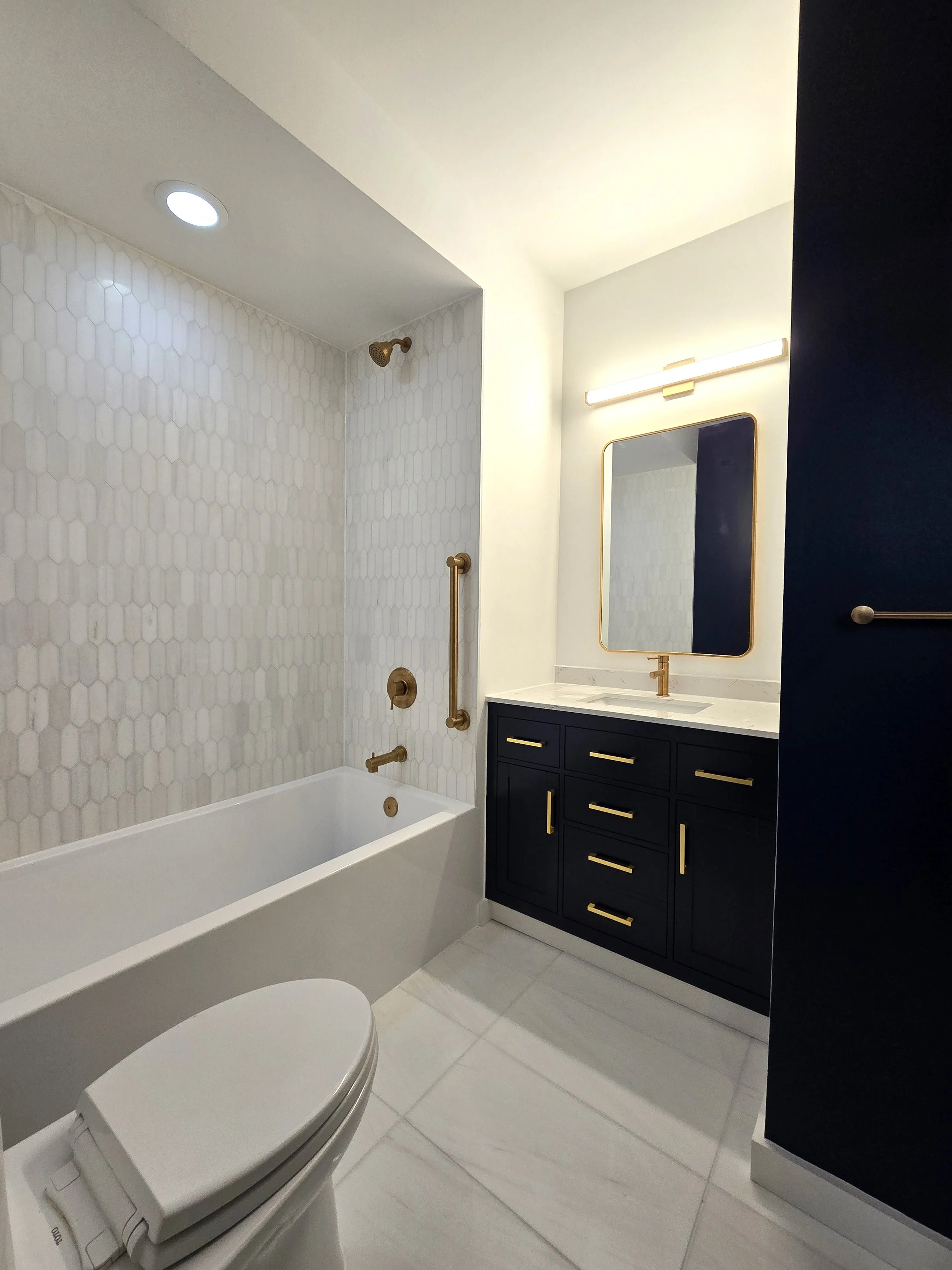 Modern bathroom renovation featuring a white bathtub with brass fixtures, a black vanity cabinet with brass handles, a marble countertop, a rectangular mirror with a brass frame, a stylish light fixture above the mirror, white hexagonal marble tile.
