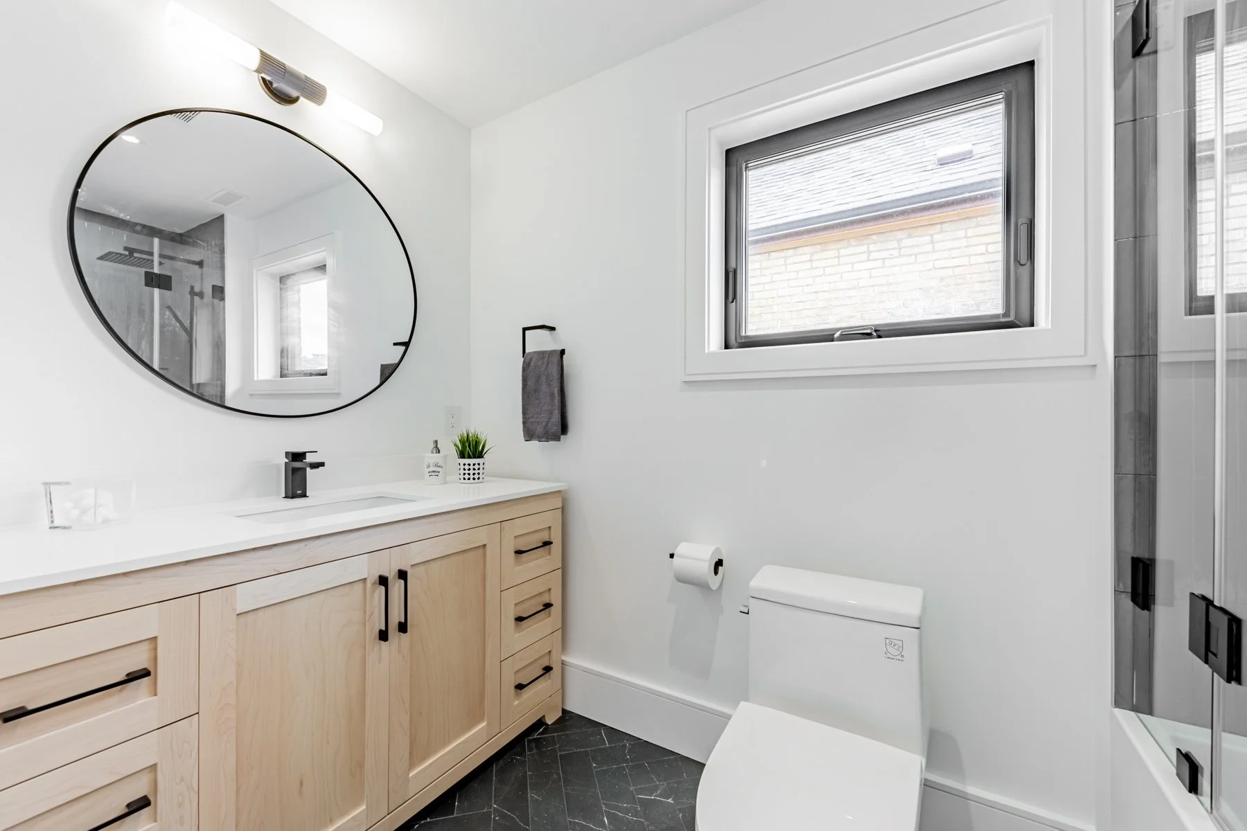 Modern bathroom renovation with a light wood vanity, black hardware, large oval mirror, and a window with a view of brick exterior wall.
