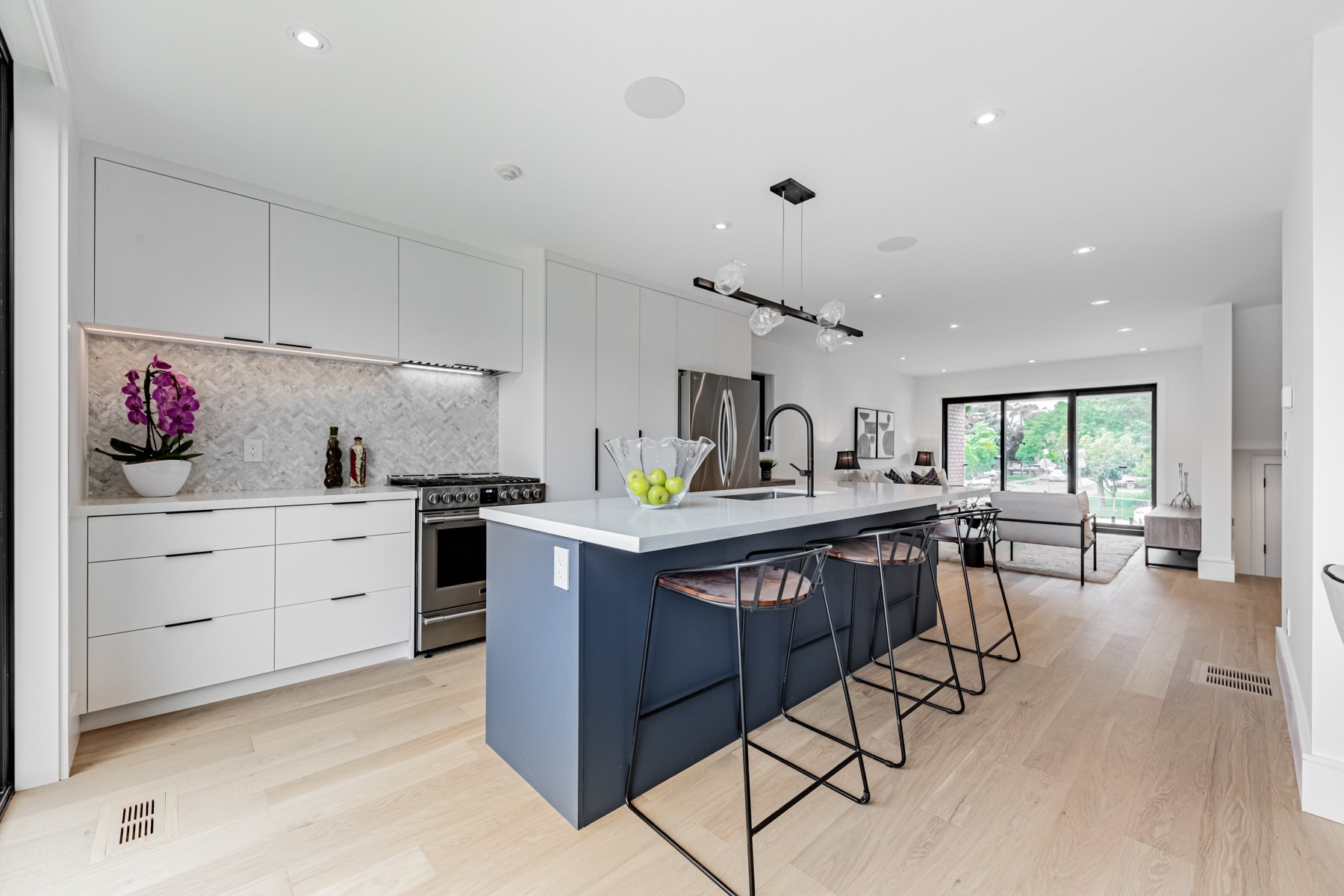 Modern open-concept kitchen renovation with white cabinetry, a blue island with a sink, black barstools, stainless steel appliances, and a living area with large windows.