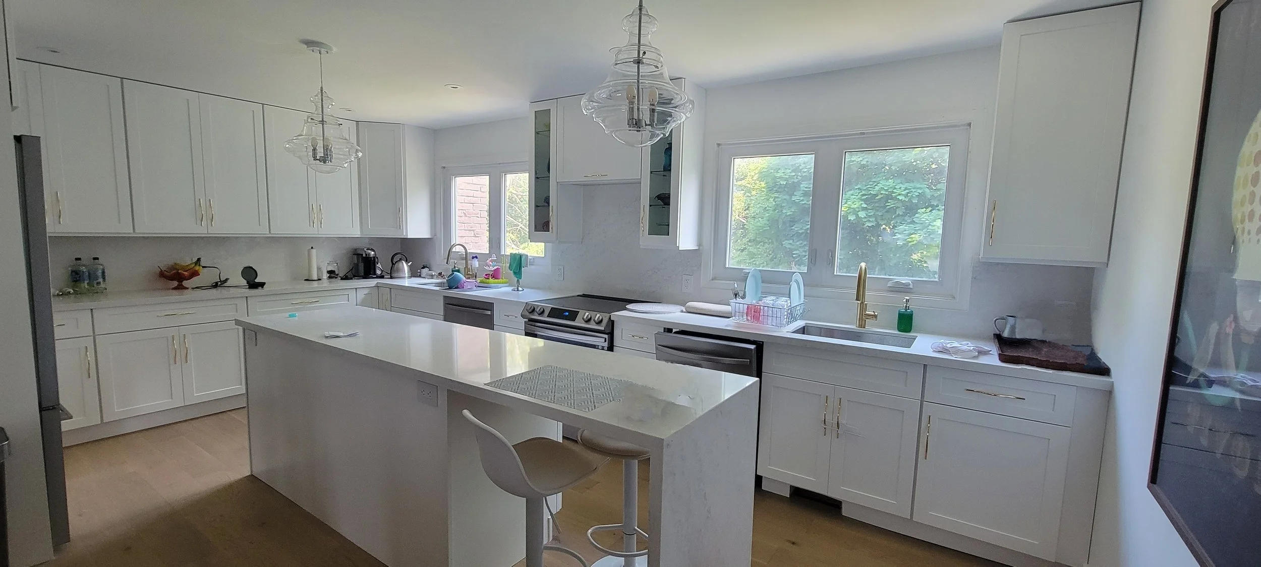 Bright, modern kitchen renovation with white cabinets, large windows, and an island with a barstool. Kitchen utensils and appliances are on the counters, with a view of green trees outside.