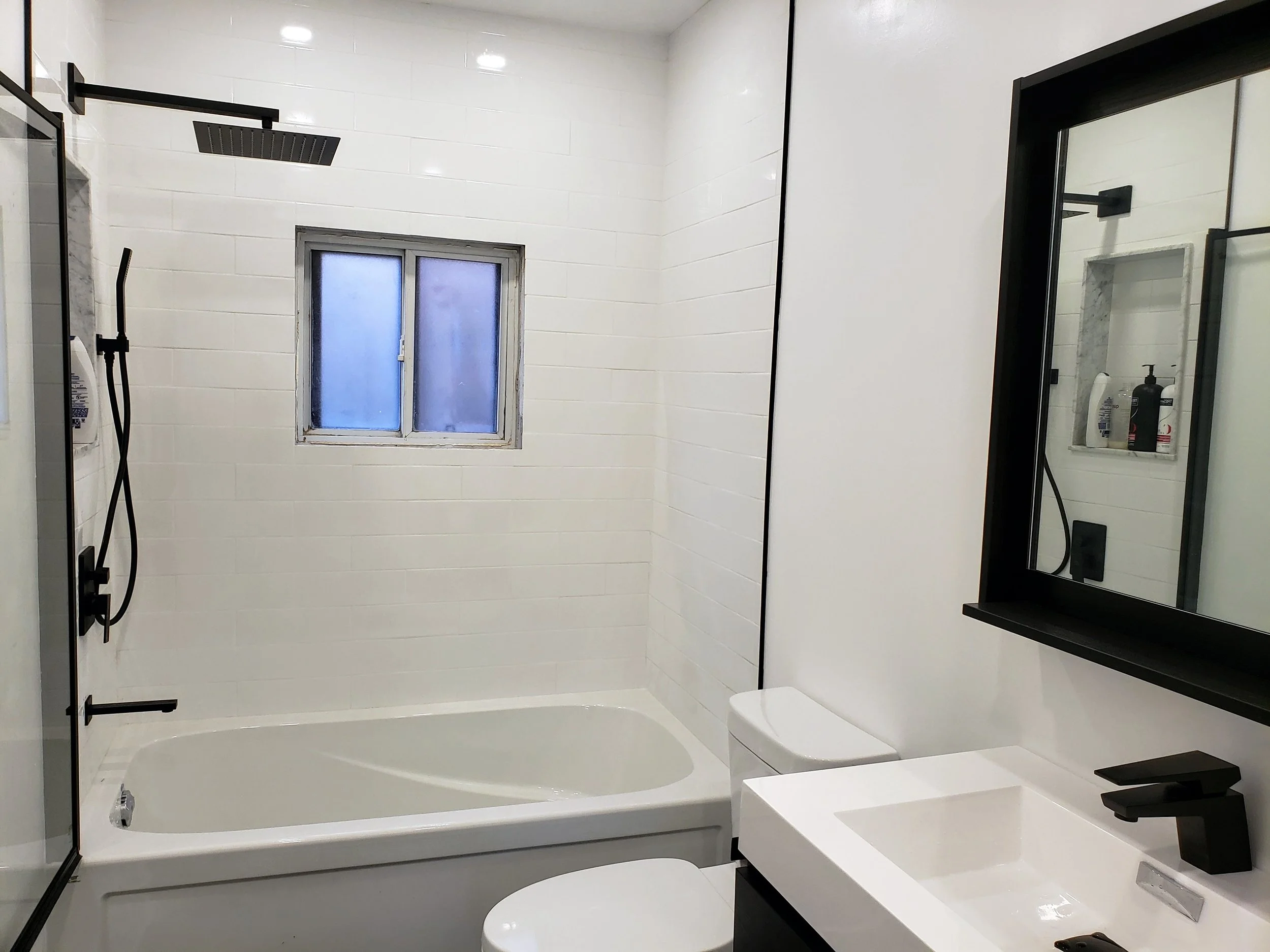 A modern bathroom renovation with a bathtub, a window, a black-framed mirror, a white sink with a black faucet, and a shower area with a rainfall showerhead and handheld shower, all with white tiled walls.