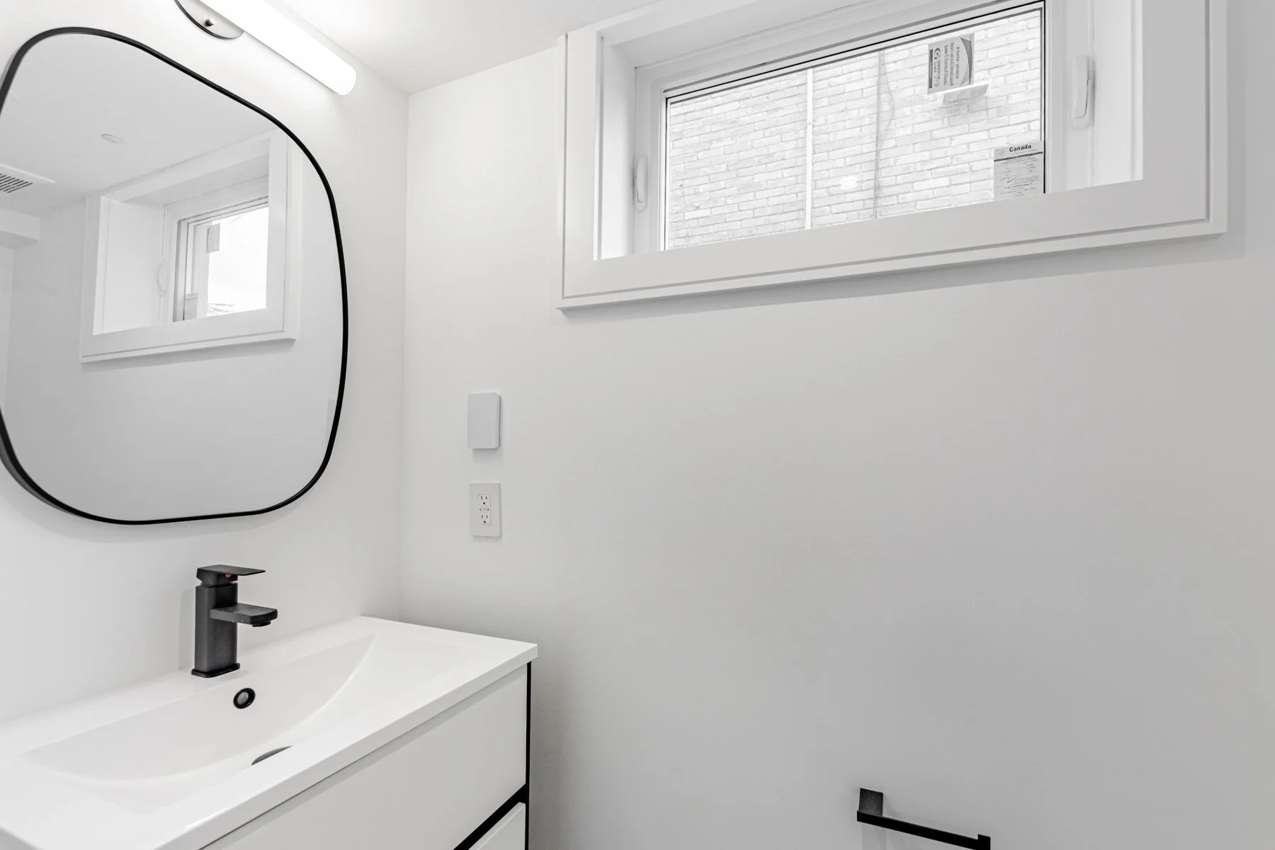 A modern bathroom renovation with a white sink, black faucet, and an irregularly shaped mirror on a white wall. There's a window showing part of a brick wall outside and bathroom fixtures, cords, and outlets on the wall.