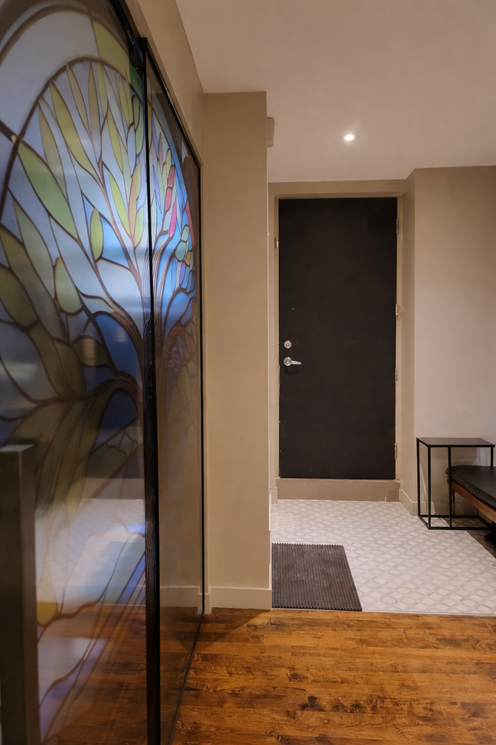 Indoor hallway with a black front door, a small black bench with a cushion, a decorative glass panel on the left, a dark doormat, and wood and patterned tile flooring.